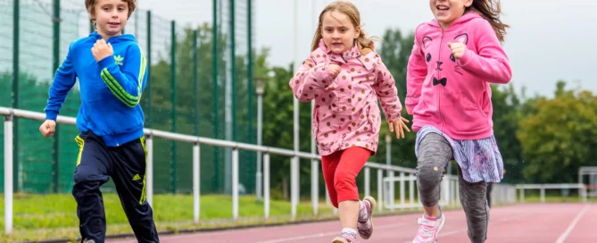 Drei Kinder in Sportkleidung laufen an einem bewölkten Tag auf einer Laufbahn im Freien, im Hintergrund sind ein Zaun und Bäume zu sehen.