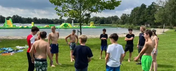 Eine Gruppe von Jungen in Badehosen steht im Kreis um einen Fußball auf einer Wiese in der Nähe eines Sees, im Hintergrund sind aufblasbare Spielgeräte zu sehen.