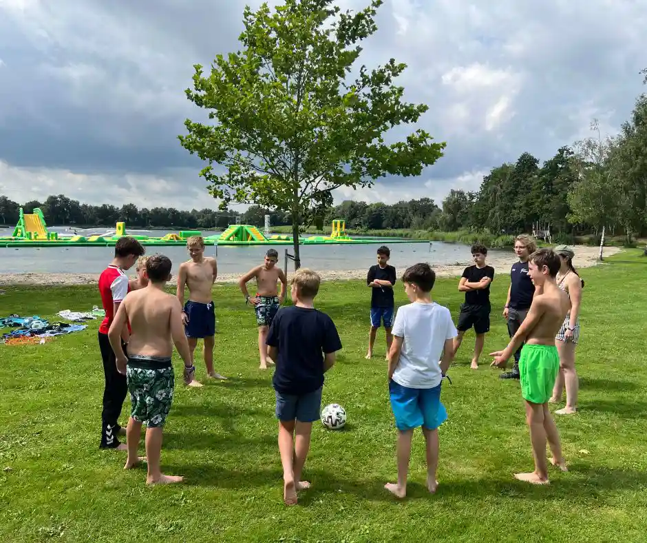 Eine Gruppe von Jungen in Badehosen steht im Kreis um einen Fußball auf einer Wiese in der Nähe eines Sees, im Hintergrund sind aufblasbare Spielgeräte zu sehen.