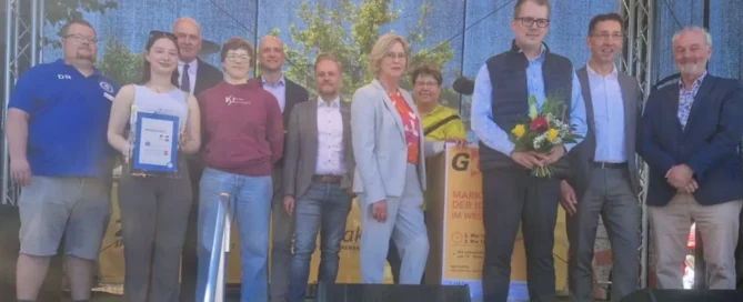 Eine Gruppe von Menschen posiert auf einer Bühne im Freien; eine Person hält eine Urkunde in der Hand, eine andere einen Blumenstrauß.