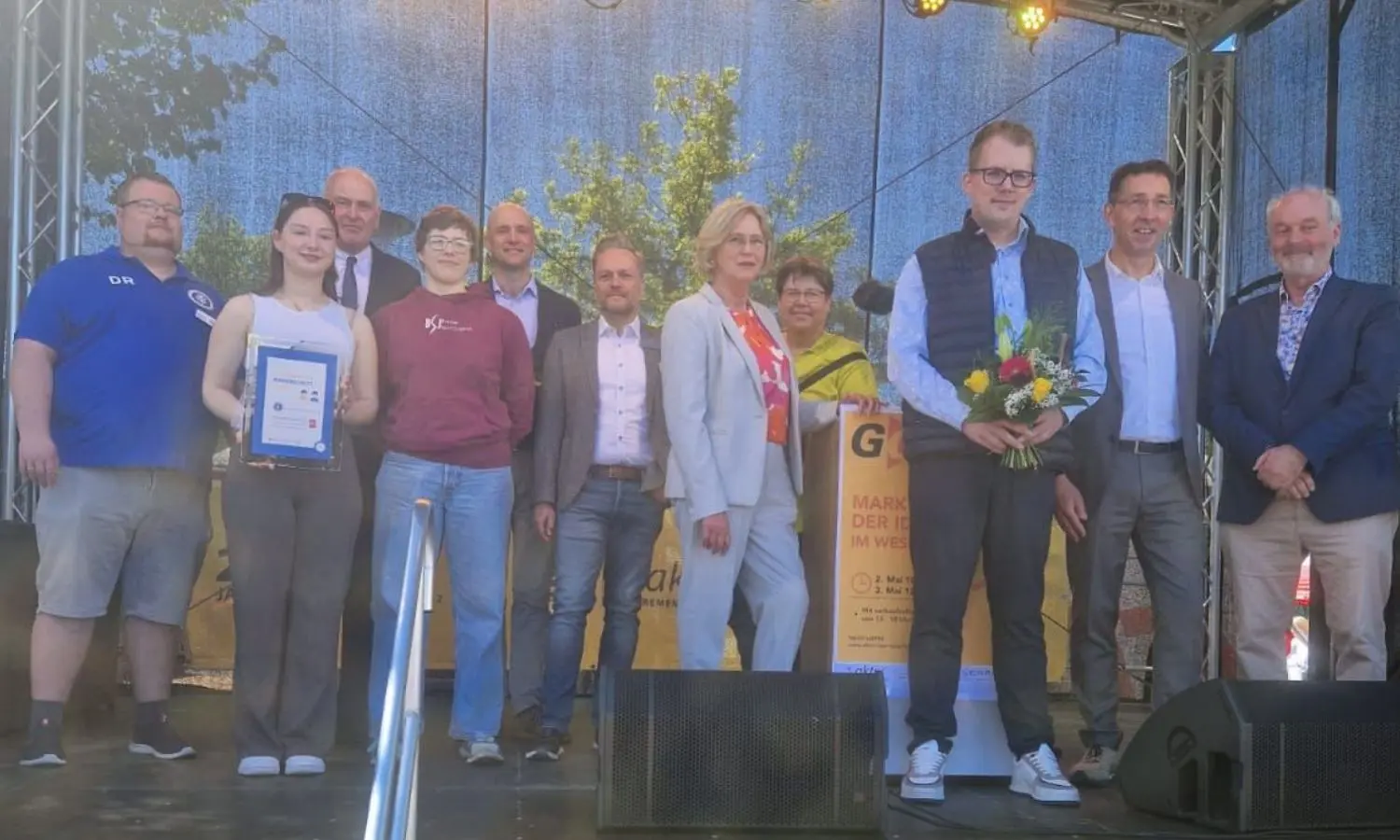 Eine Gruppe von Menschen posiert auf einer Bühne im Freien; eine Person hält eine Urkunde in der Hand, eine andere einen Blumenstrauß.