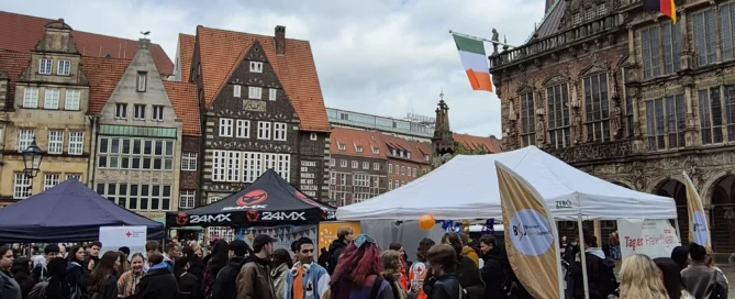 Auf einem Stadtplatz in einer europäischen Stadt (Bremen) versammelt sich eine Menschenmenge bei einer Veranstaltung unter freiem Himmel mit Zelten; unter dem bewölkten Himmel sind historische Gebäude und Flaggen zu sehen.