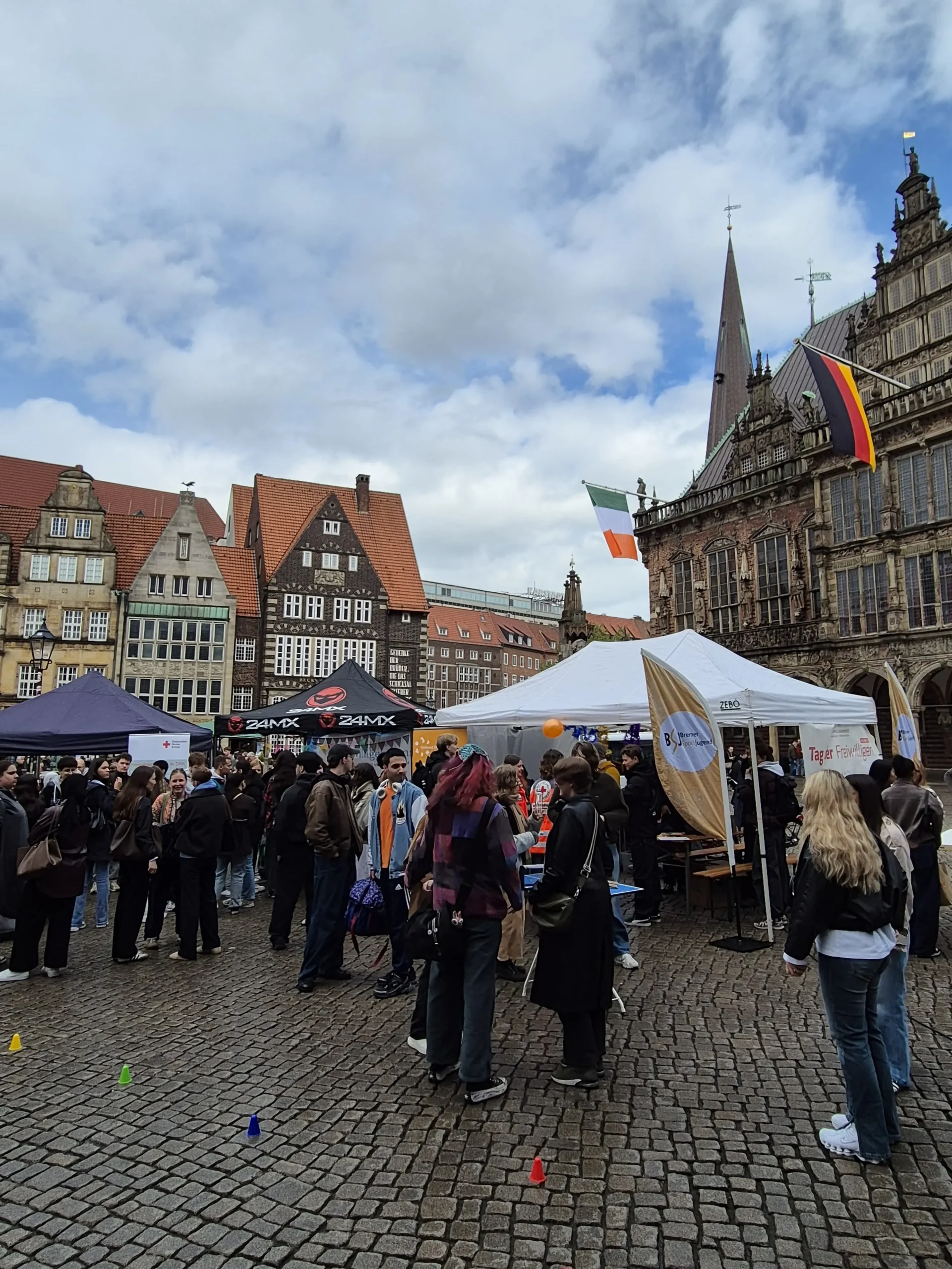 Auf einem Stadtplatz in einer europäischen Stadt (Bremen) versammelt sich eine Menschenmenge bei einer Veranstaltung unter freiem Himmel mit Zelten; unter dem bewölkten Himmel sind historische Gebäude und Flaggen zu sehen.