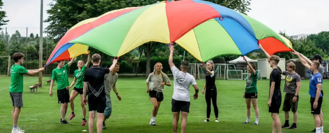 Eine Gruppe von Menschen steht im Freien im Kreis auf einer Wiese und hält einen großen, bunten Fallschirm hoch