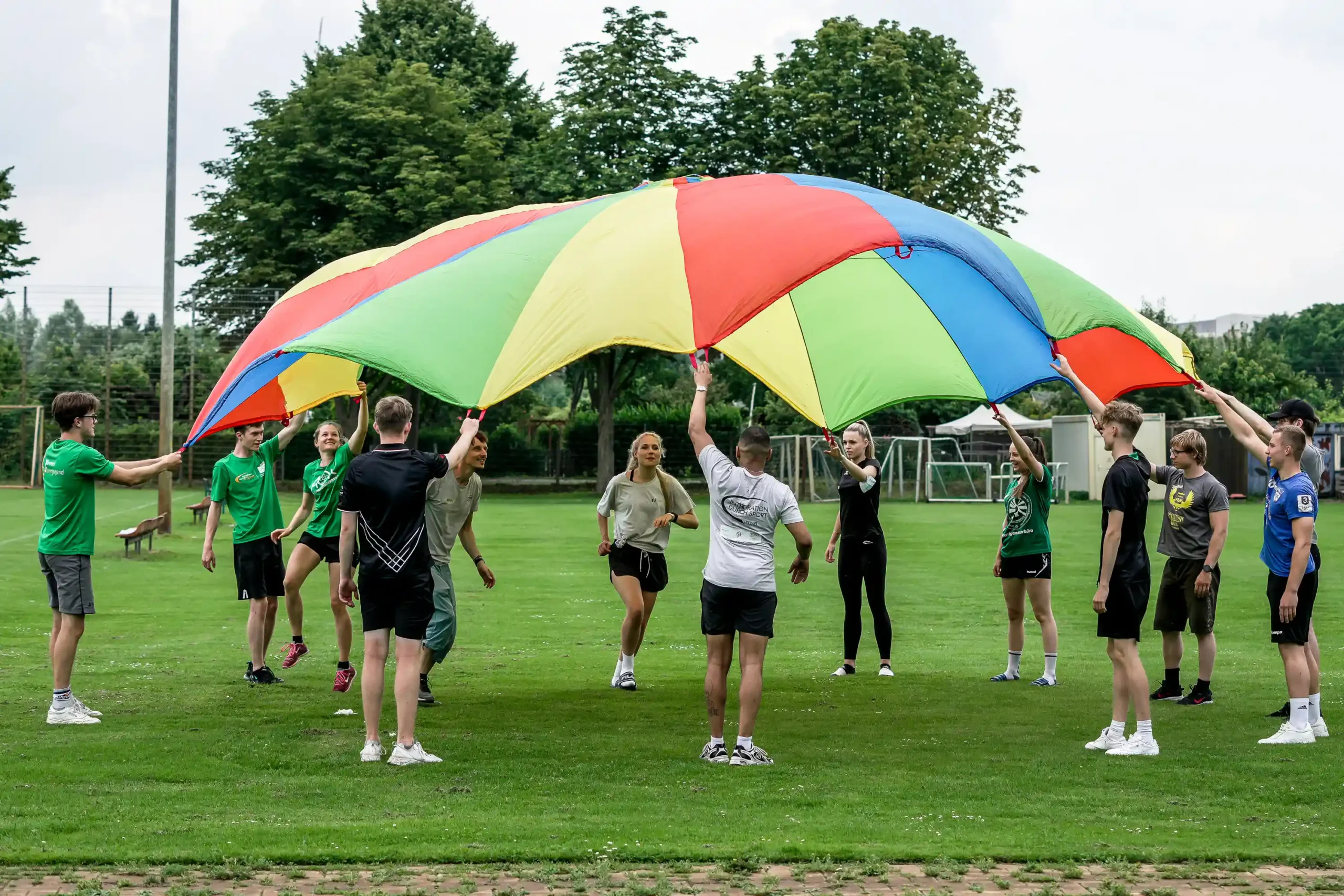 Eine Gruppe von Menschen steht im Freien im Kreis auf einer Wiese und hält einen großen, bunten Fallschirm hoch
