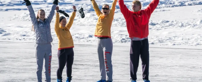 Vier Menschen, die an einem sonnigen Wintertag im Freien Eislaufen, die Arme in die Höhe strecken und lächeln; sie tragen legere Winterkleidung und blaue Schlittschuhe.