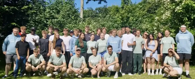 Eine Gruppe von etwa 30 Personen posiert für ein Foto auf einer Wiese, im Hintergrund sind Bäume und der blaue Himmel zu sehen. Die meisten stehen, einige knien vorne, und alle scheinen sich im Freien zu befinden.