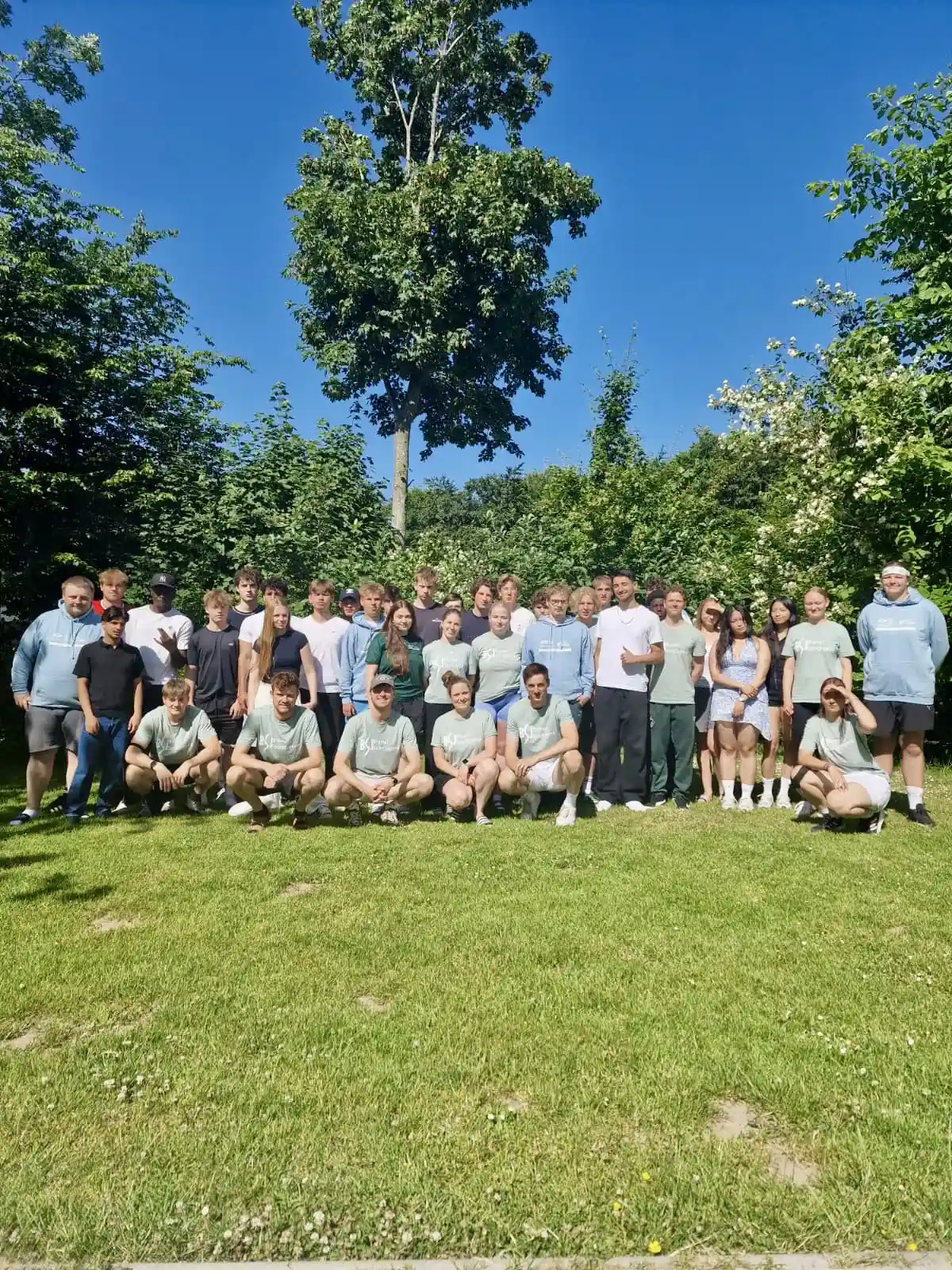 Eine Gruppe von etwa 30 Personen posiert für ein Foto auf einer Wiese, im Hintergrund sind Bäume und der blaue Himmel zu sehen. Die meisten stehen, einige knien vorne, und alle scheinen sich im Freien zu befinden.