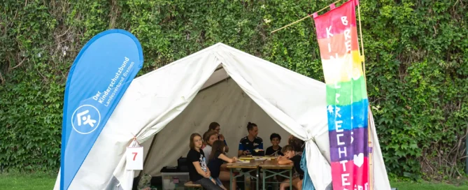 Kinder sitzen an Tischen in einem weißen Zelt, das im Freien aufgestellt ist; am Eingang hängen ein blaues Transparent und eine bunte Flagge. Im Hintergrund ist grünes Laub zu sehen.
