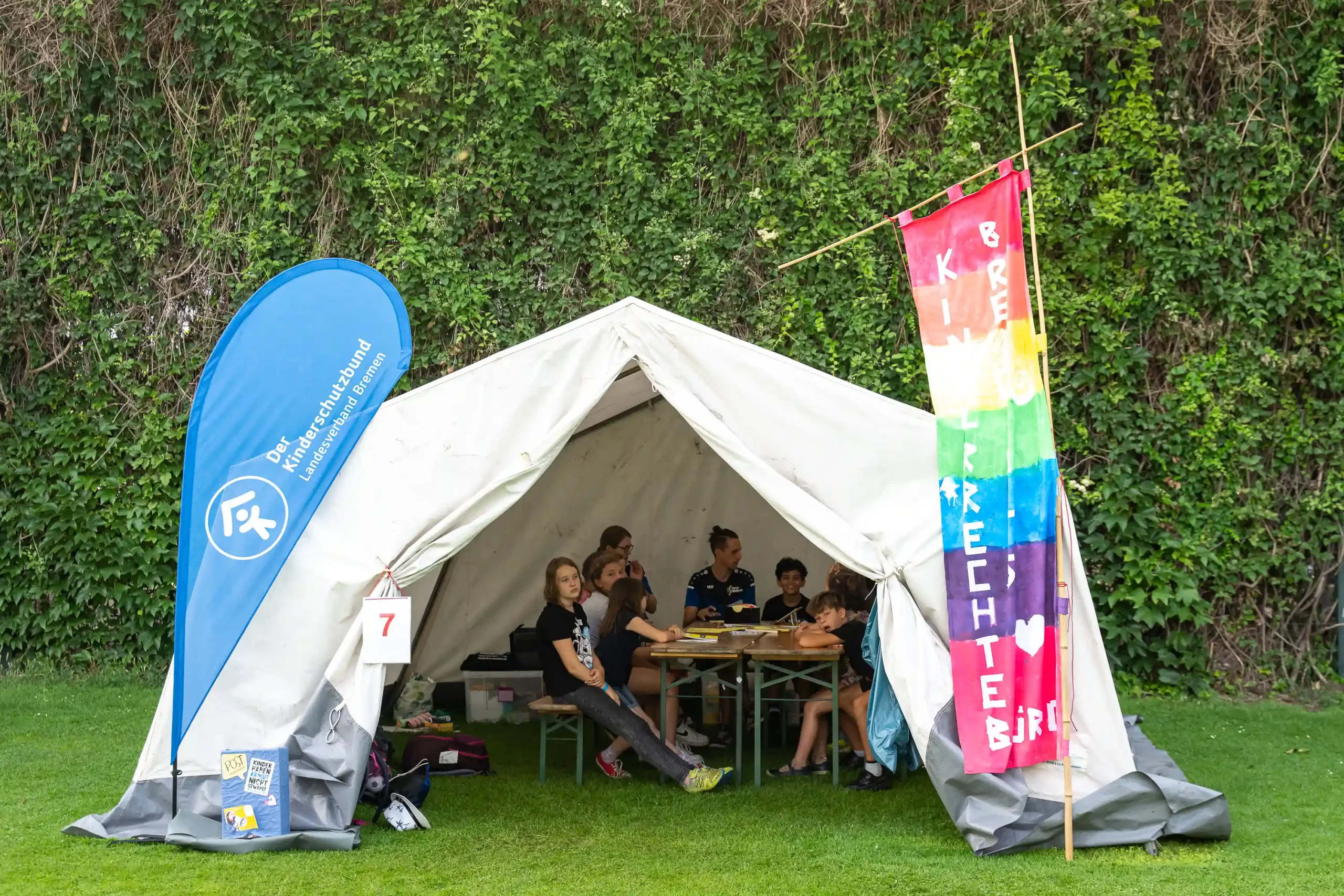 Kinder sitzen an Tischen in einem weißen Zelt, das im Freien aufgestellt ist; am Eingang hängen ein blaues Transparent und eine bunte Flagge. Im Hintergrund ist grünes Laub zu sehen.