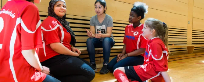 Ein Trainer sitzt auf dem Turnhallenbank und unterhält sich während einer Mannschaftsbesprechung mit vier jungen Fußballspielern in roten Trikots.