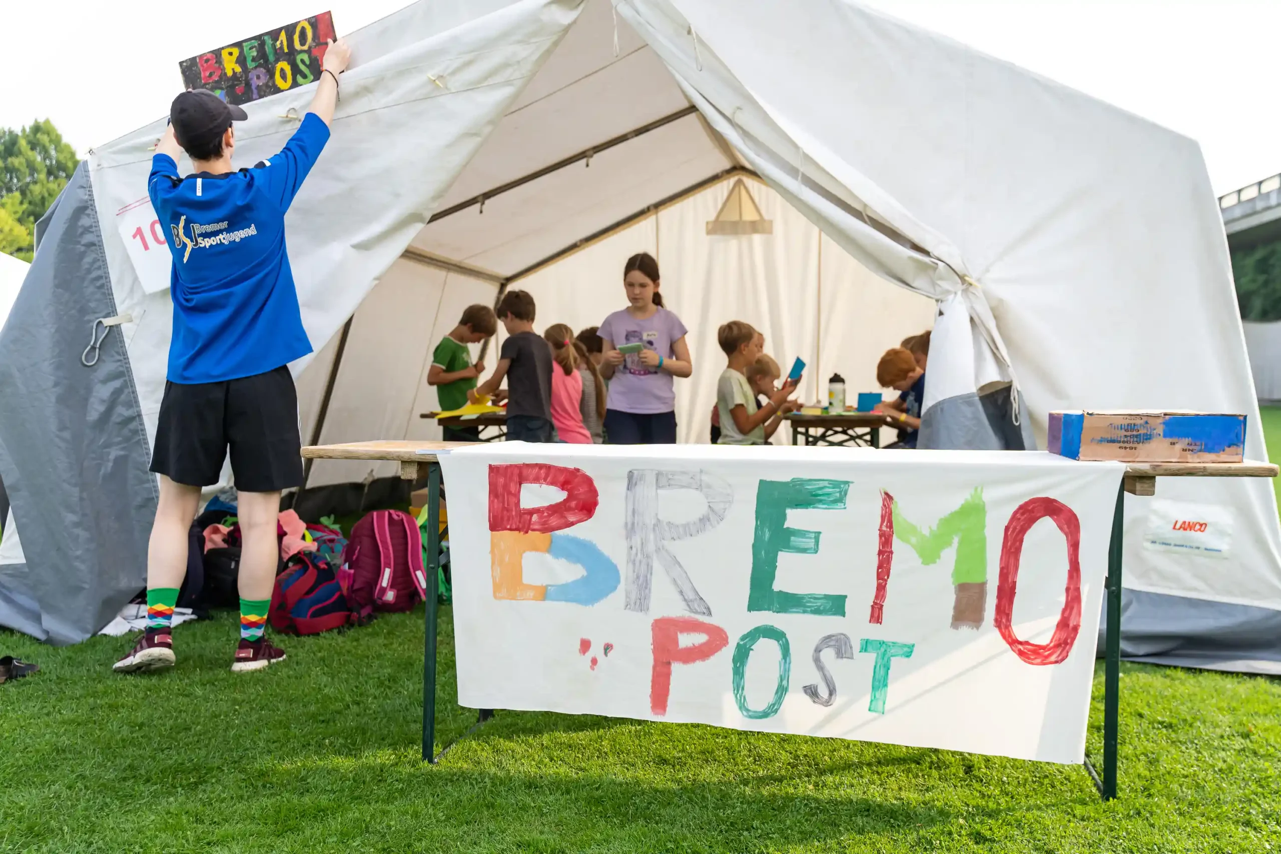 Eine Person hängt ein buntes Schild mit der Aufschrift „BREMO POST“ an ein Zelt, während Kinder drinnen an Tischen verschiedenen Aktivitäten nachgehen. Ein passendes Schild steht draußen auf einem Tisch.