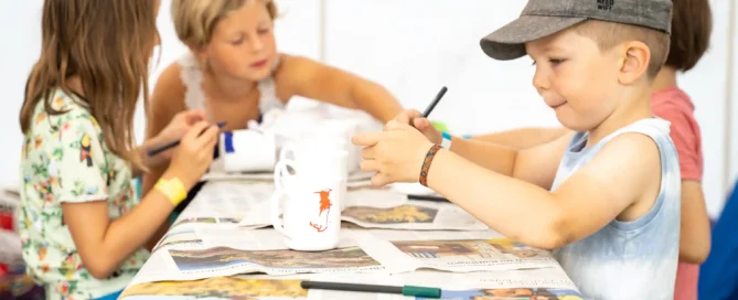 Vier Kinder sitzen an einem mit Zeitungen bedeckten Tisch und malen und verzieren Gegenstände. Die Atmosphäre wirkt ungezwungen und kreativ, auf dem Tisch liegen Malutensilien.