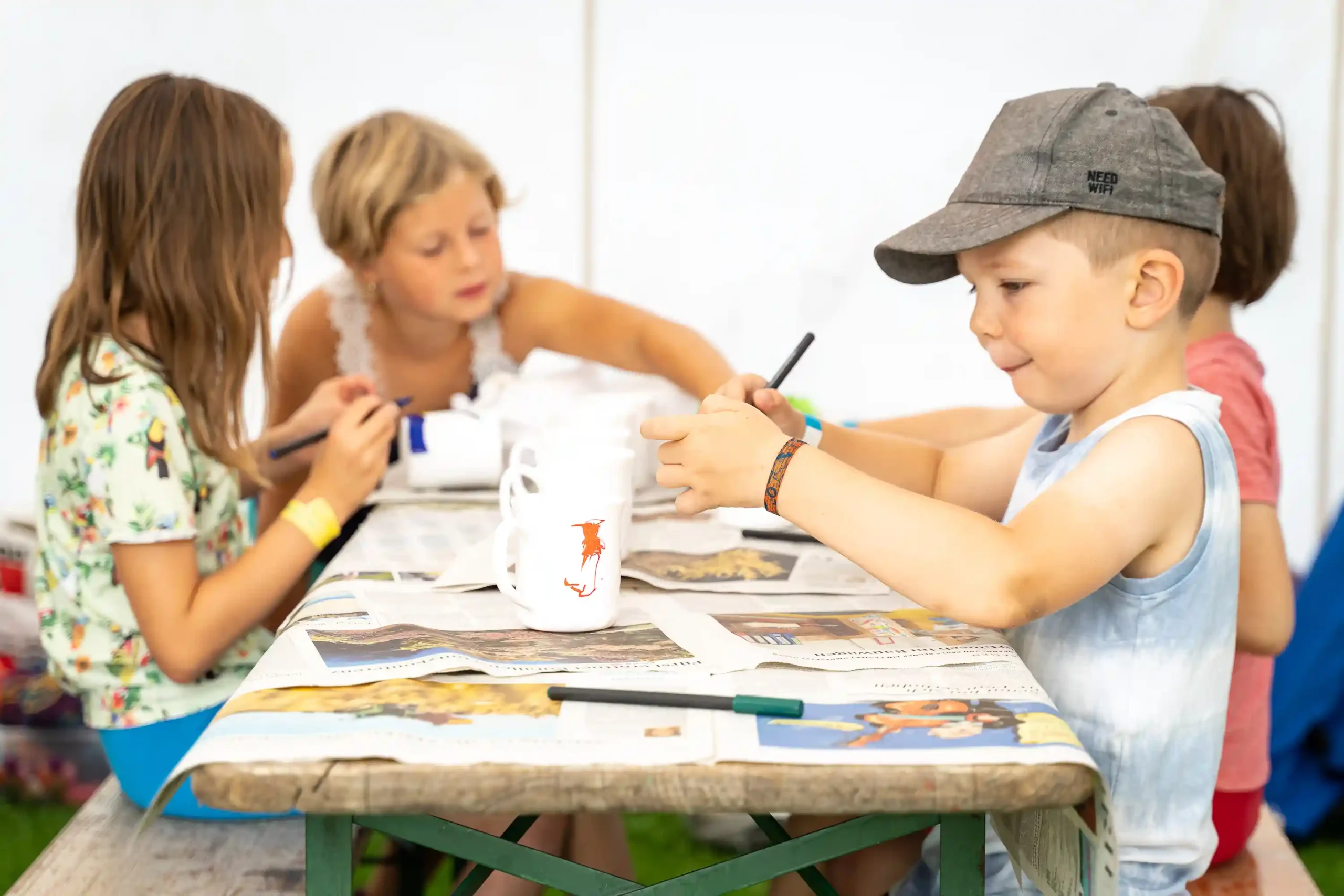 Vier Kinder sitzen an einem mit Zeitungen bedeckten Tisch und malen und verzieren Gegenstände. Die Atmosphäre wirkt ungezwungen und kreativ, auf dem Tisch liegen Malutensilien.