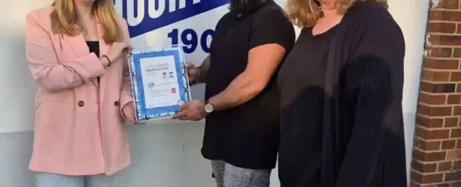 Drei Personen stehen vor einem Schild mit der Aufschrift „TUS Huchting“; der Mann in der Mitte hält eine Urkunde in der Hand, während die beiden Frauen lächeln.