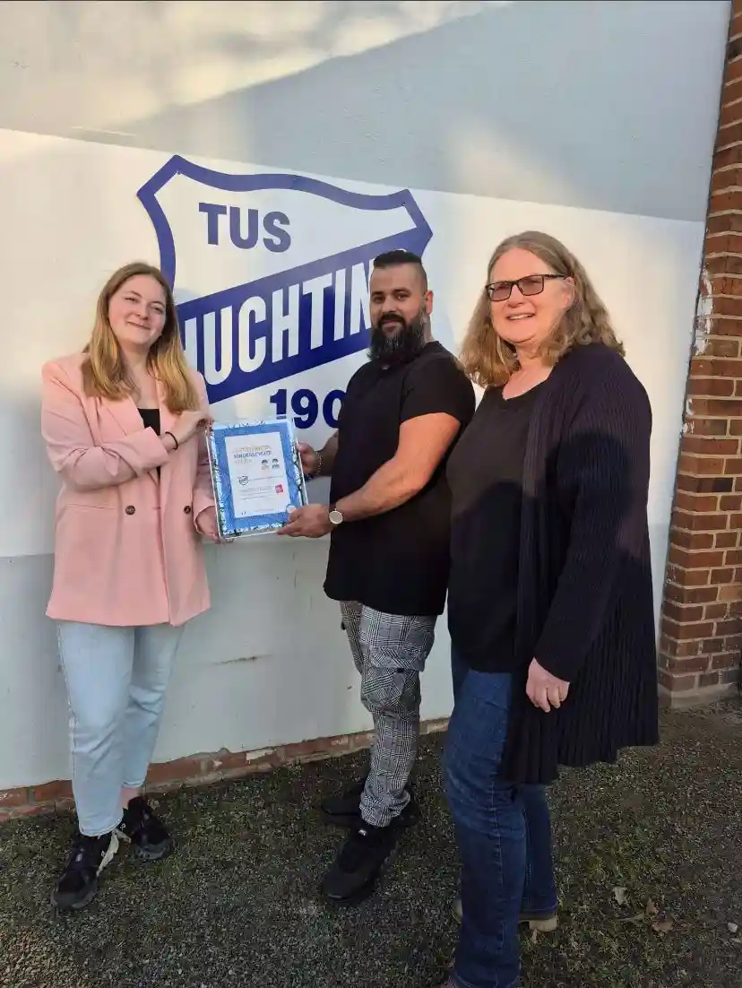 Drei Personen stehen vor einem Schild mit der Aufschrift „TUS Huchting“; der Mann in der Mitte hält eine Urkunde in der Hand, während die beiden Frauen lächeln.