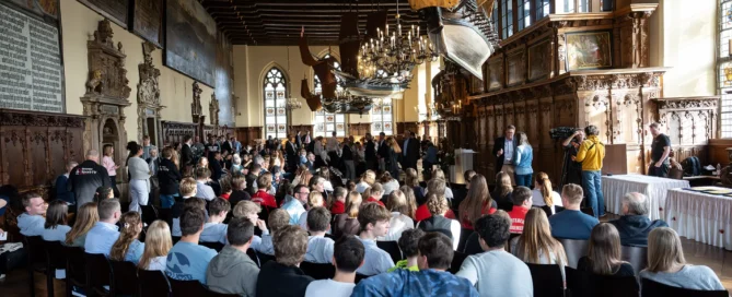 Eine große Gruppe von Menschen sitzt in Stuhlreihen in einem prächtigen Saal mit Holzwänden, Kronleuchtern und Schiffsmodellen, die von der Decke hängen.