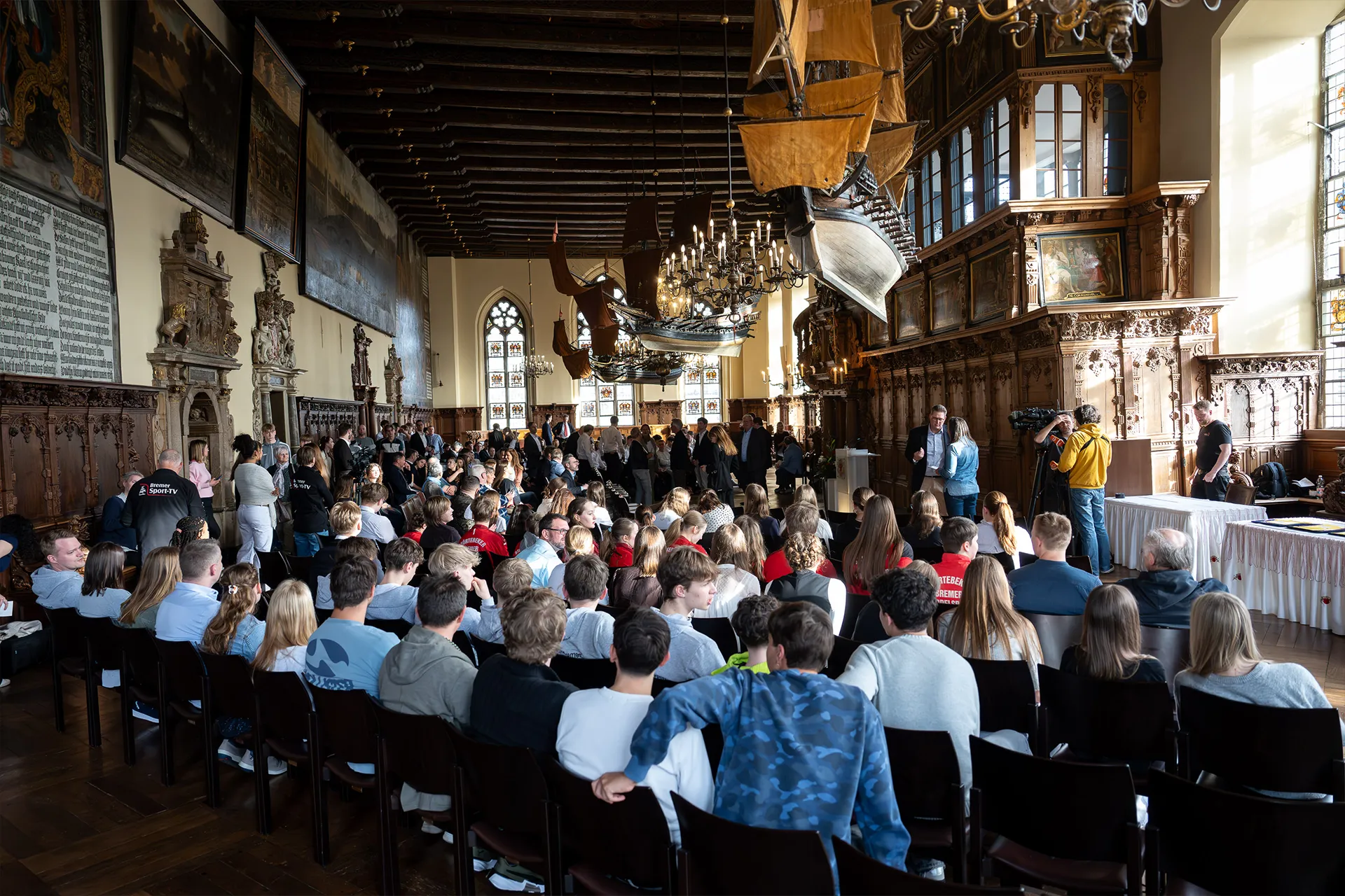Eine große Gruppe von Menschen sitzt in Stuhlreihen in einem prächtigen Saal mit Holzwänden, Kronleuchtern und Schiffsmodellen, die von der Decke hängen.