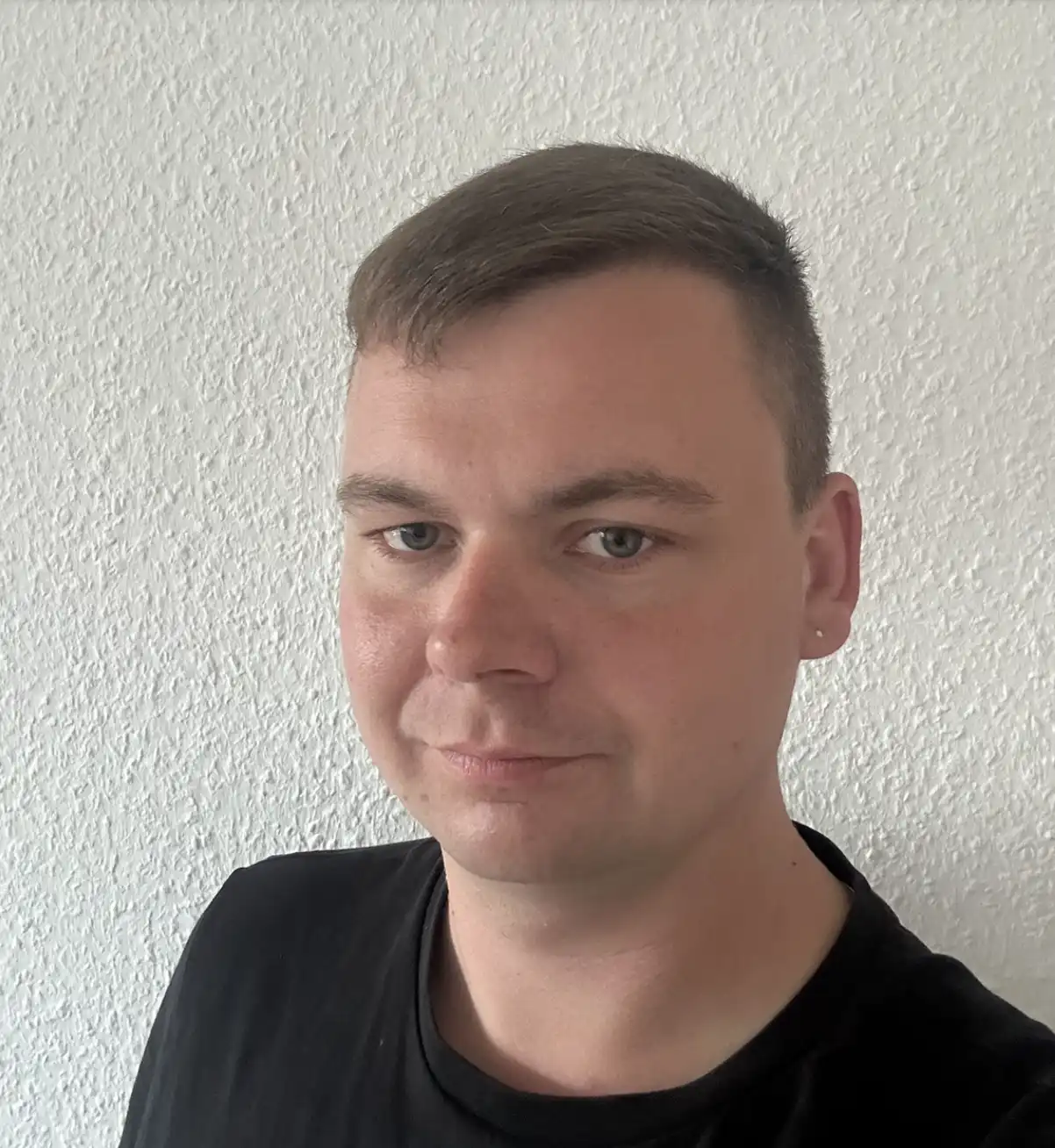 BSJ_Stimmen_Sebastain Eine Person mit kurzen braunen Haaren und heller Haut, die ein schwarzes Hemd trägt, steht vor einer strukturierten weißen Wand und blickt in die Kamera.