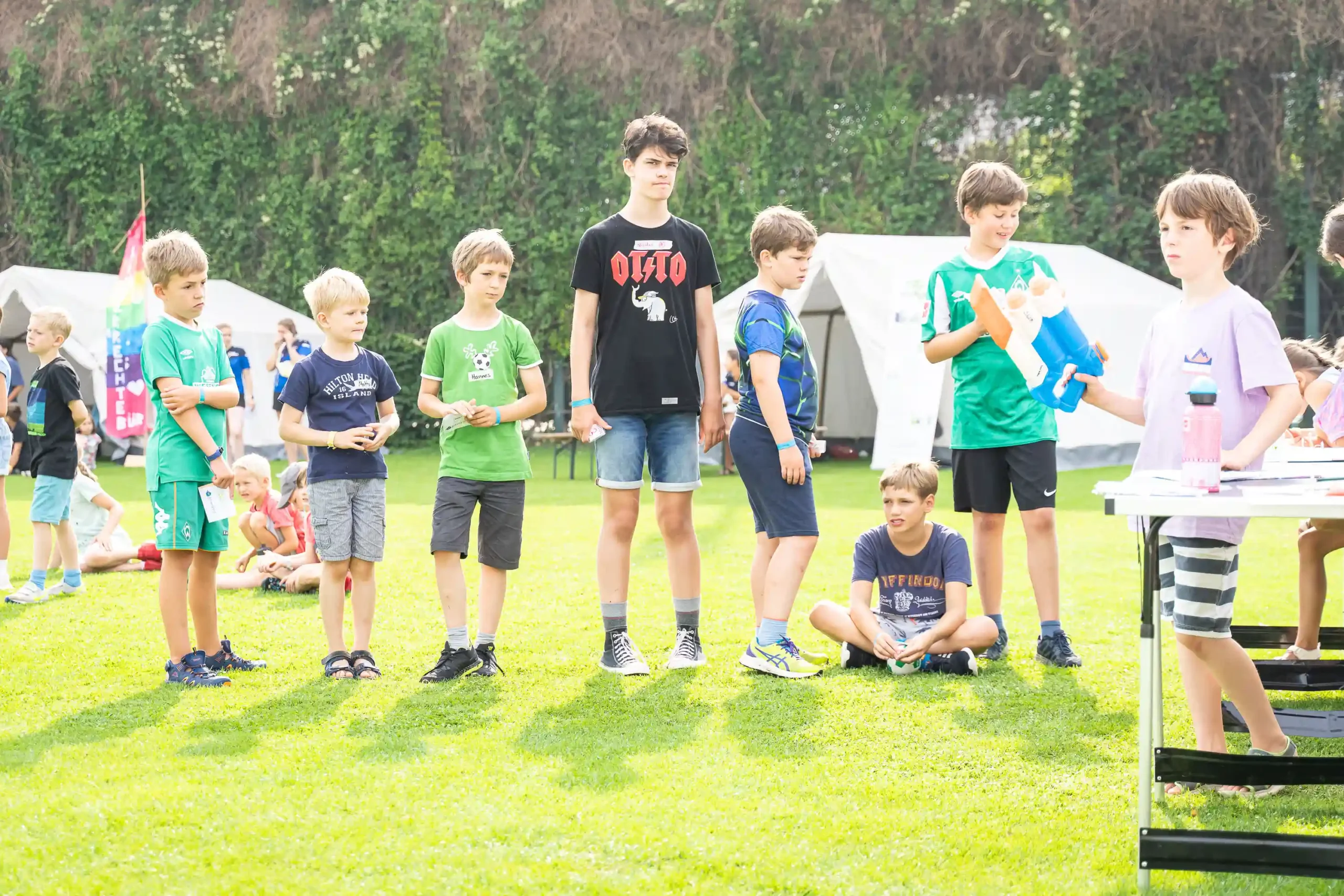Eine Gruppe von Jungen steht in einer Reihe auf einer Wiese, im Hintergrund sind Zelte und Bäume zu sehen. Ein Junge sitzt an einem Tisch und hält einen blauen Gegenstand in der Hand, während die anderen zuschauen oder darauf warten, dass sie an die Reihe kommen.