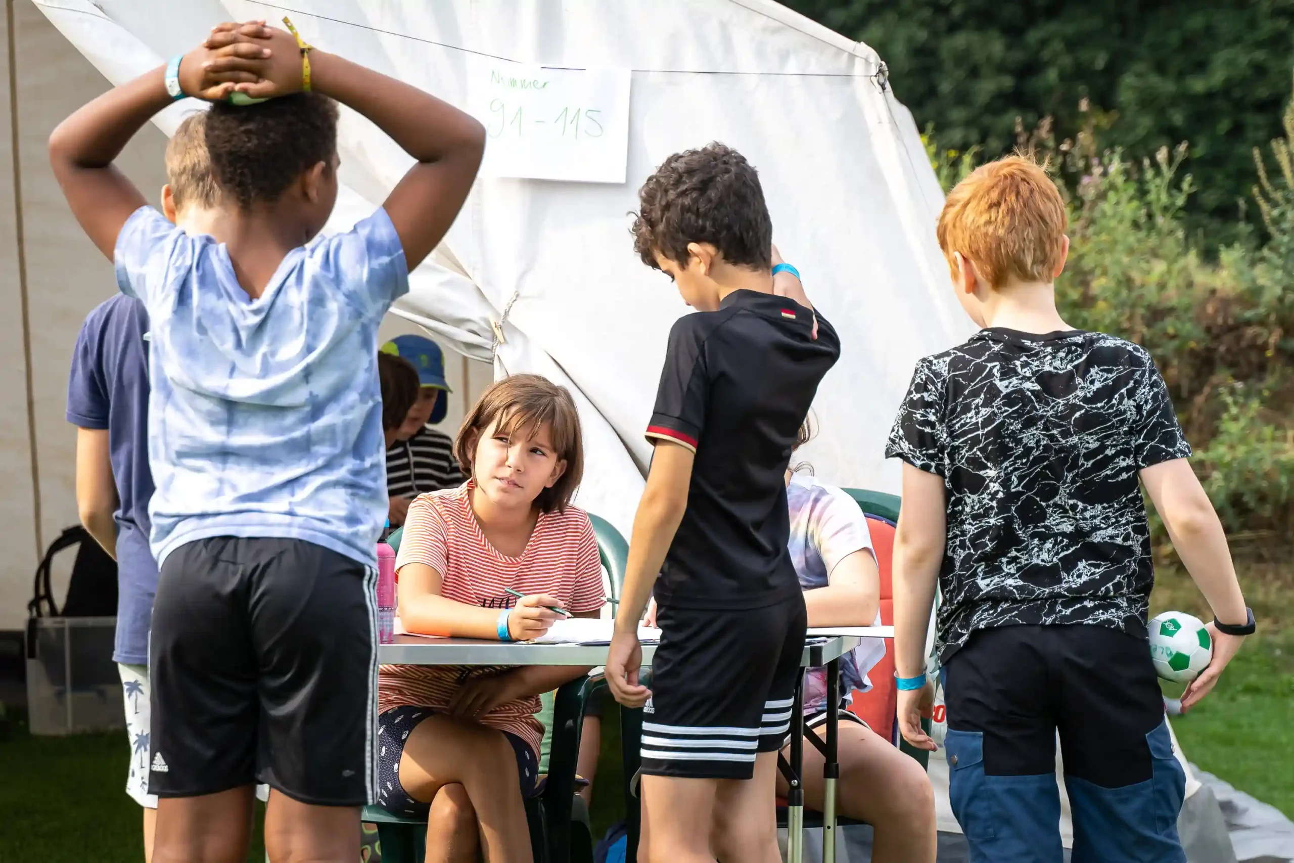 Vier Kinder stehen vor einem Zelt in der Nähe eines Tisches und unterhalten sich, während ein weiteres Kind dort sitzt und auf ein Blatt Papier schreibt. Ein Kind hält einen Fußball in der Hand. Im Hintergrund sind Bäume und Gras zu sehen.