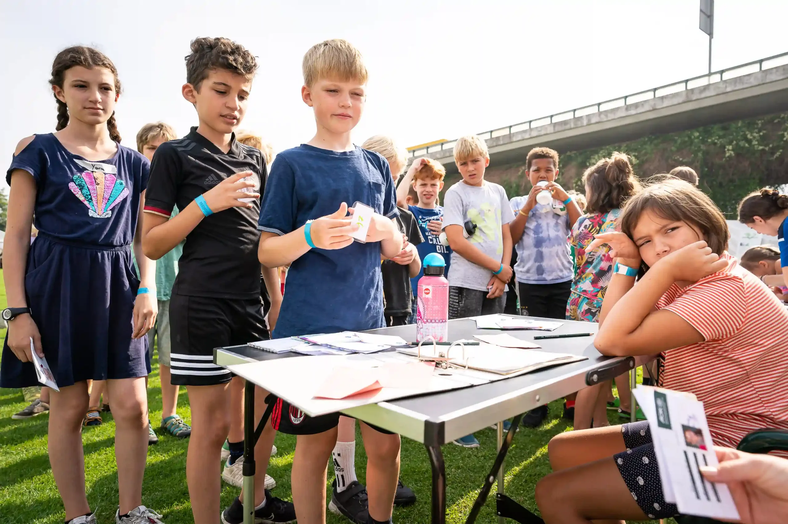 Eine Gruppe von Kindern steht und sitzt im Freien auf dem Rasen um einen Tisch herum, auf dem Papiere liegen, und ist unter freiem Himmel mit einer Aktivität beschäftigt.