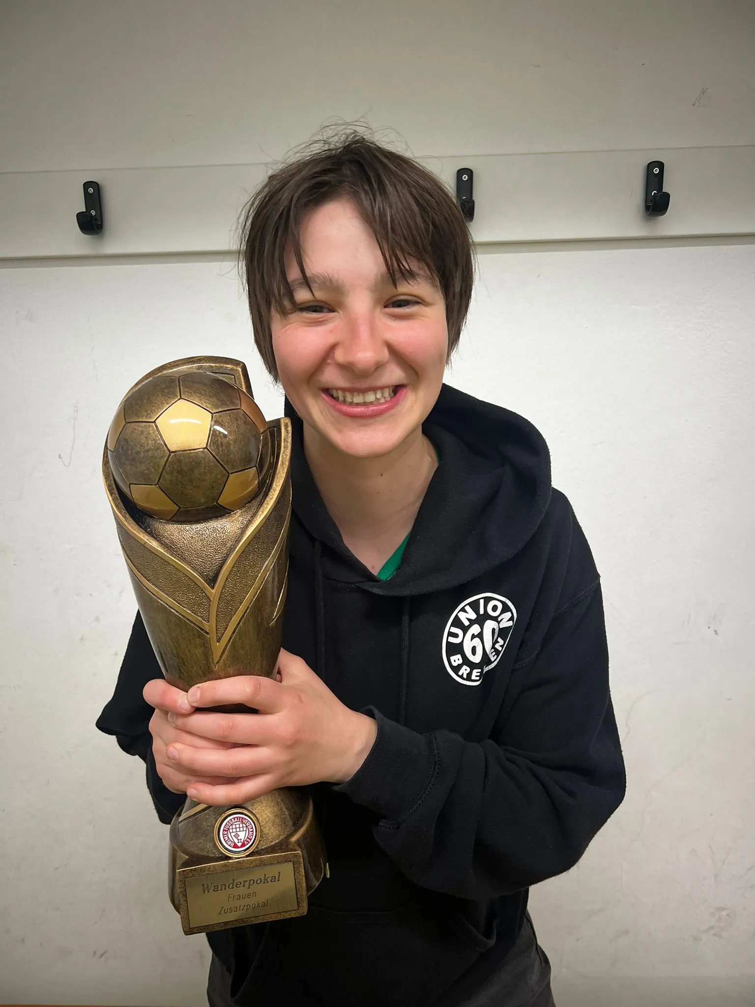 Bremer-Sportjugend_Lotte Eine lächelnde Person in einem schwarzen Kapuzenpulli hält einen großen goldenen Fußballpokal mit der Aufschrift „Wanderpokal“ vor einer weißen Wand in der Hand.