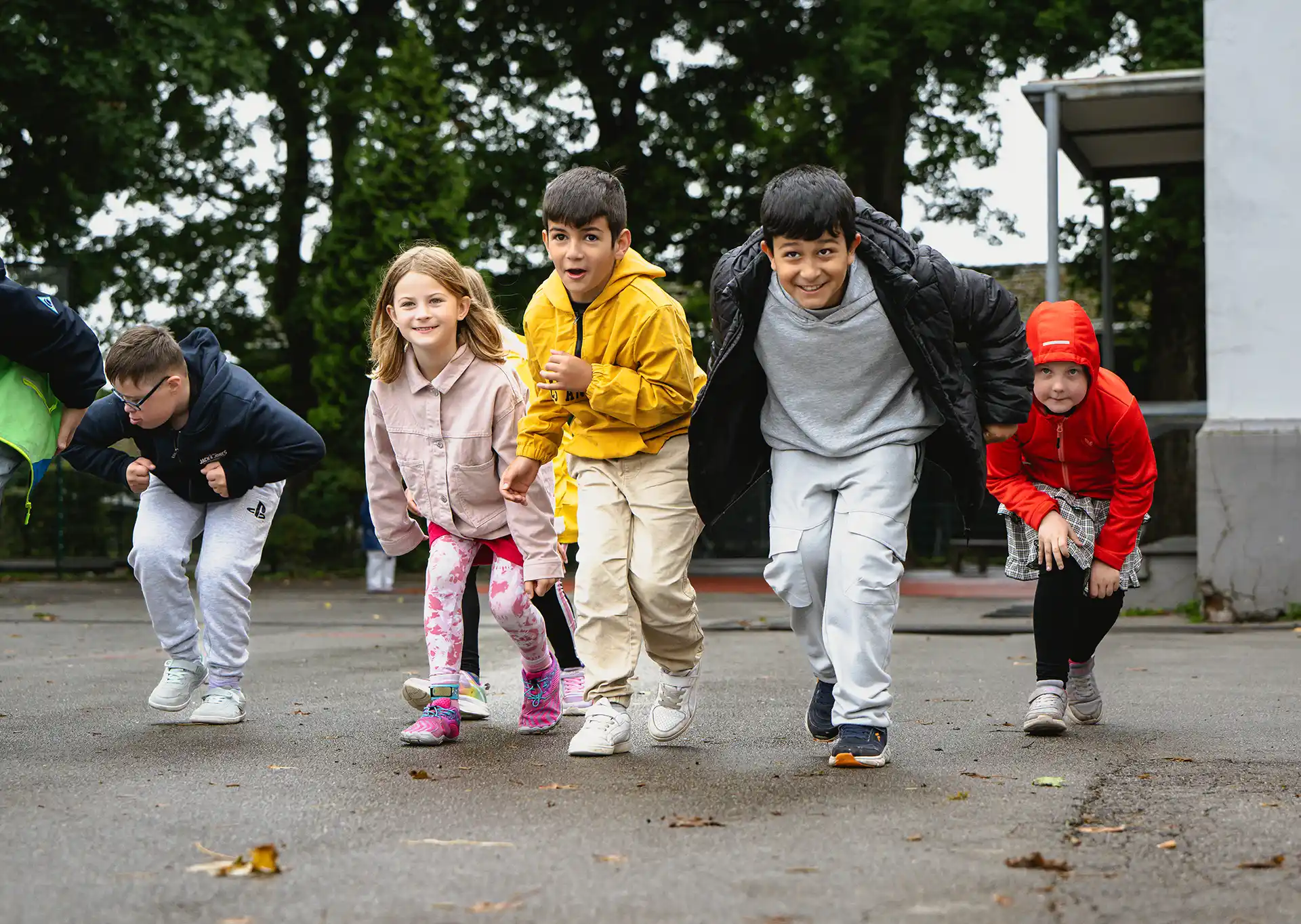 Fünf Kinder in Freizeitkleidung stehen draußen in einer Reihe und scheinen bereit zu sein, ein Rennen zu starten; im Hintergrund sind Bäume und ein Gebäude zu sehen.