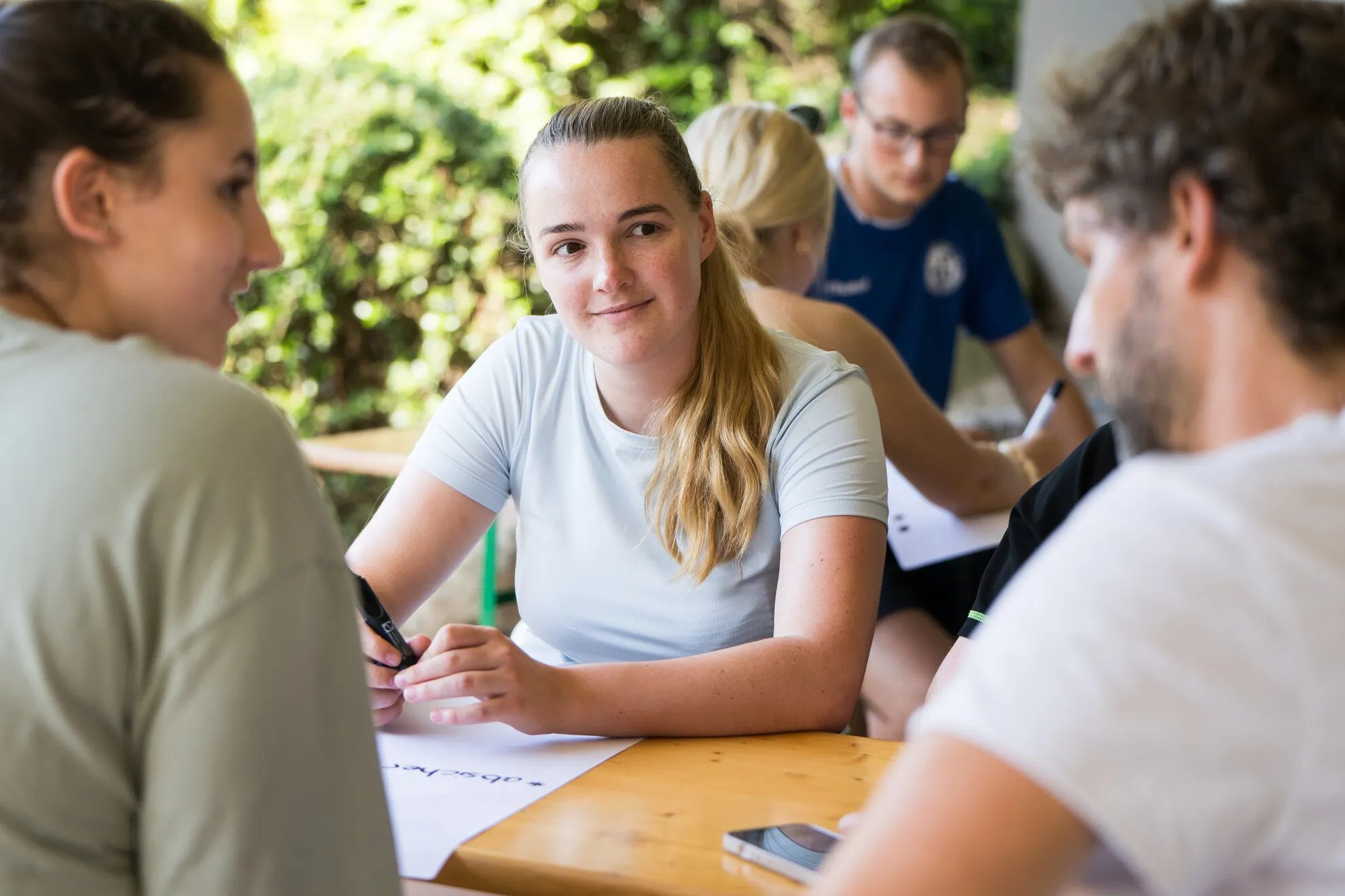 Eine Gruppe von Menschen sitzt draußen an einem Holztisch, unterhält sich und schreibt auf Papierbögen, im Hintergrund ist Grün zu sehen.