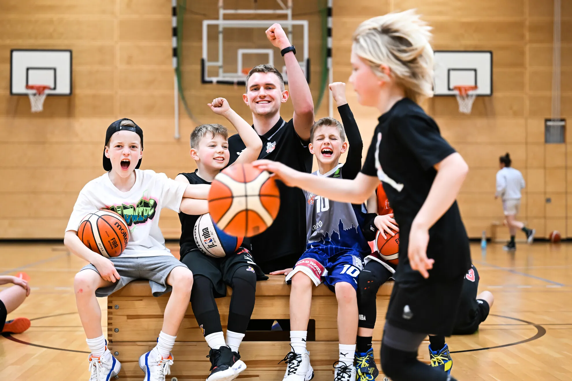 Fünf Kinder und ein Trainer sitzen auf einer Bank in einer Sporthalle, jubeln und halten Basketbälle in den Händen, während ein weiteres Kind mit einem Basketball in der Hand an ihnen vorbeirennt.