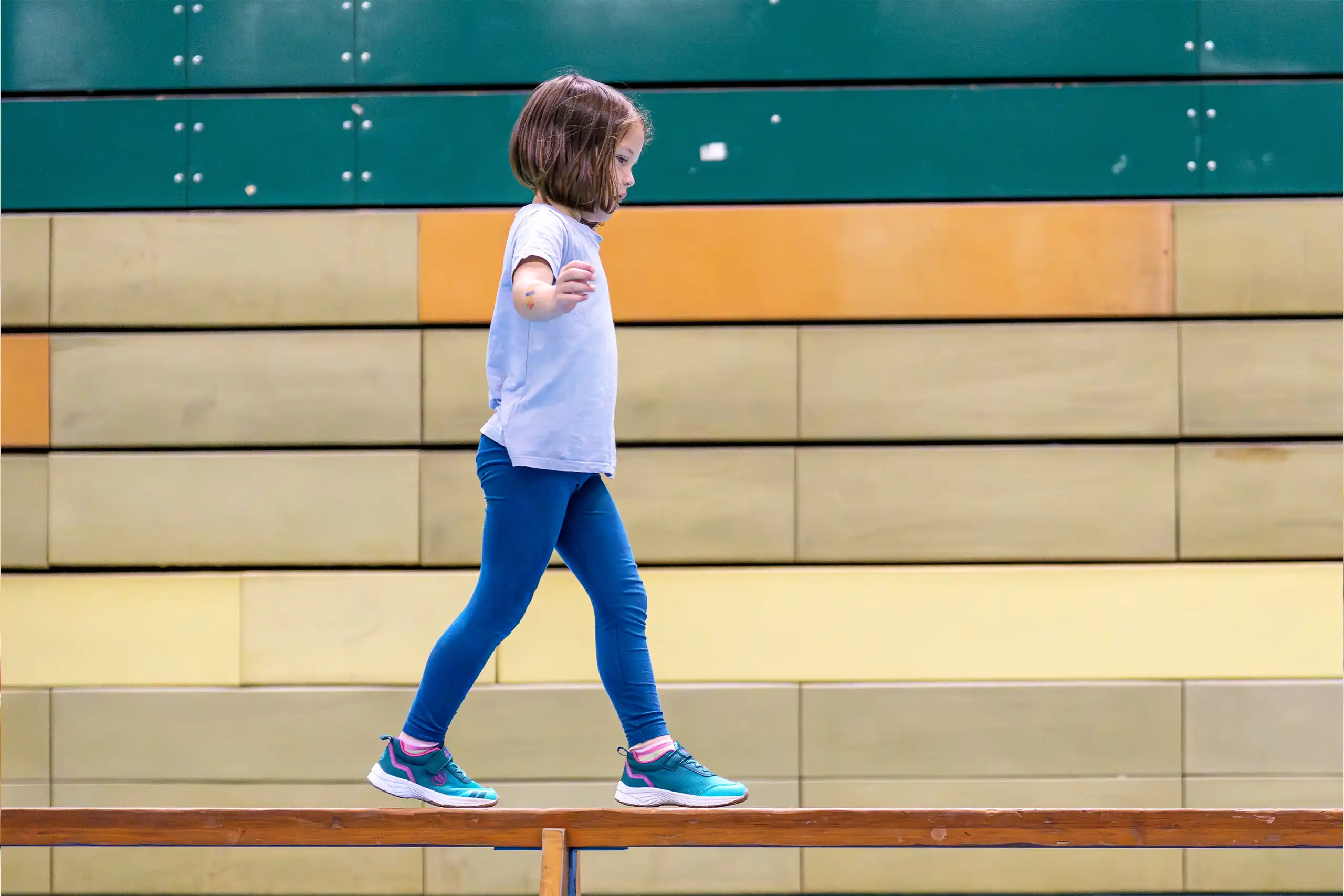 Zwei Kinder spielen drinnen ein Spiel mit Holzklötzen; das Mädchen in Rosa ist ganz in das Spiel vertieft, während der Junge in Gelb daneben steht und einen Ball in der Hand hält.