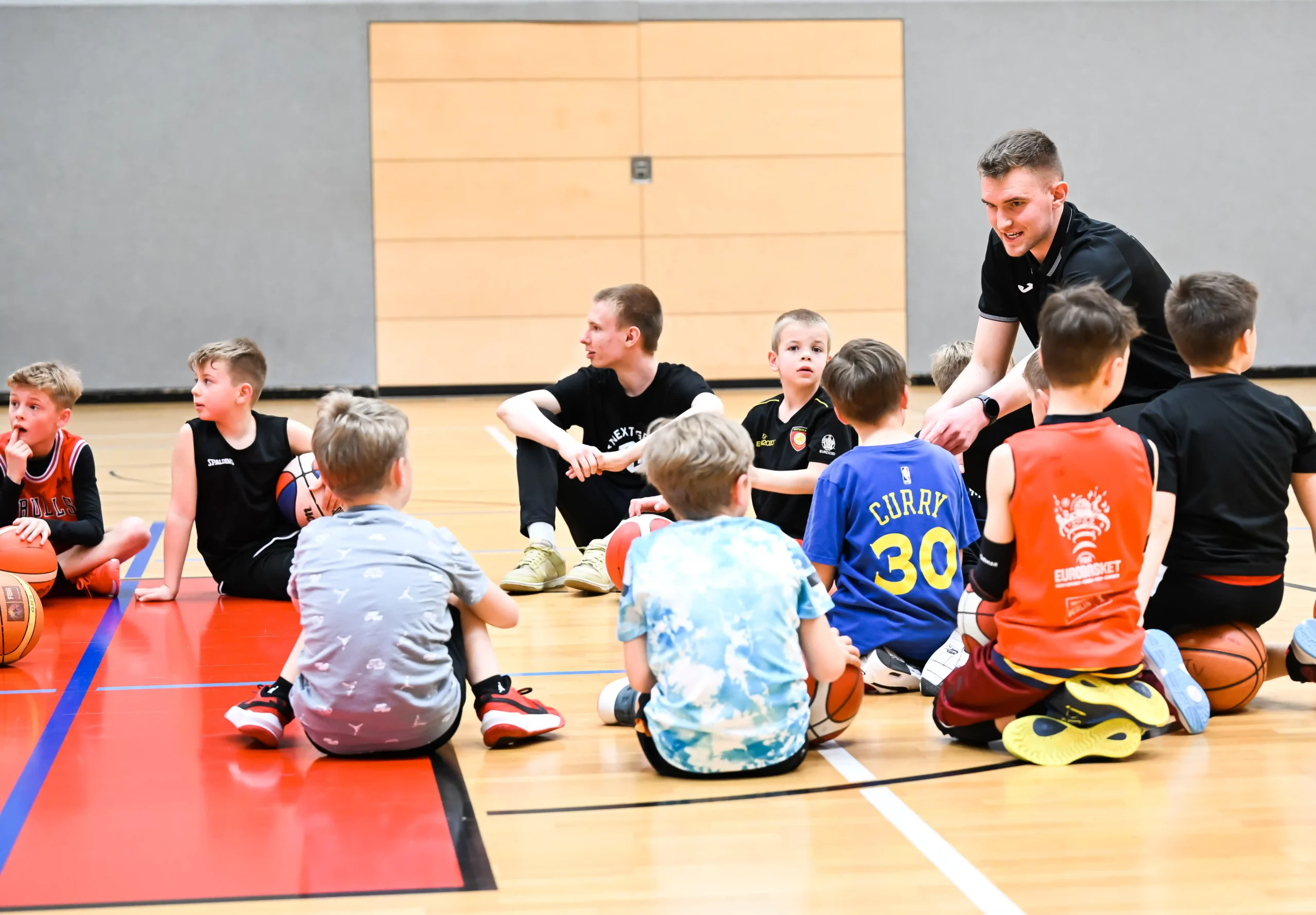 BSJ_Bremen_Basketball_Junges_Team Eine Gruppe junger Jungen sitzt mit Basketbällen auf dem Boden einer Turnhalle, während ein erwachsener Trainer während des Basketballtrainings vor ihnen kniet und mit ihnen spricht.
