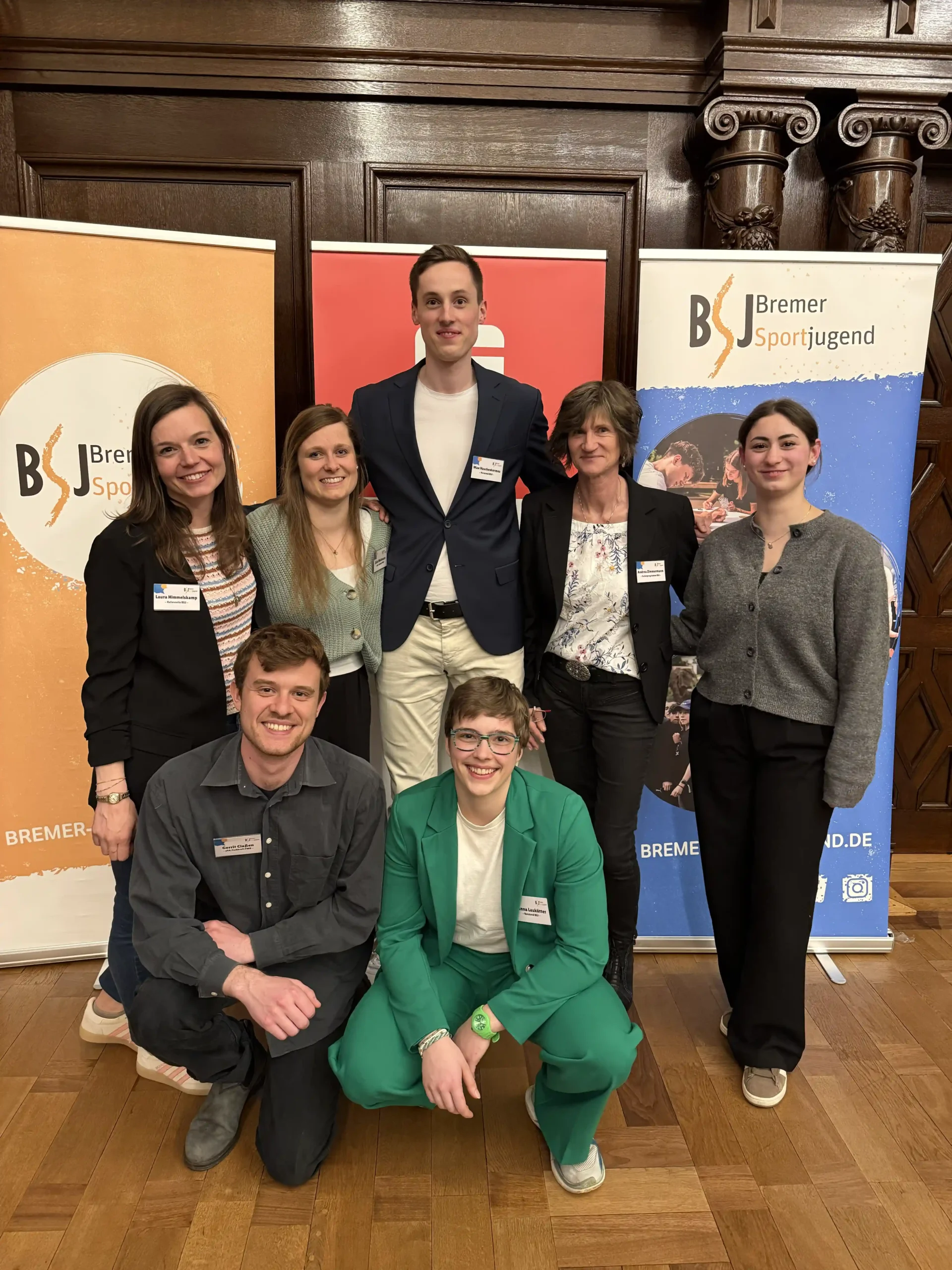 Sieben Personen posieren in einem Raum vor zwei Bannern der „Bremer Sportjugend“ und einem roten Hintergrund; sie tragen Business- und Freizeitkleidung und lächeln für ein Gruppenfoto auf einem Holzboden.