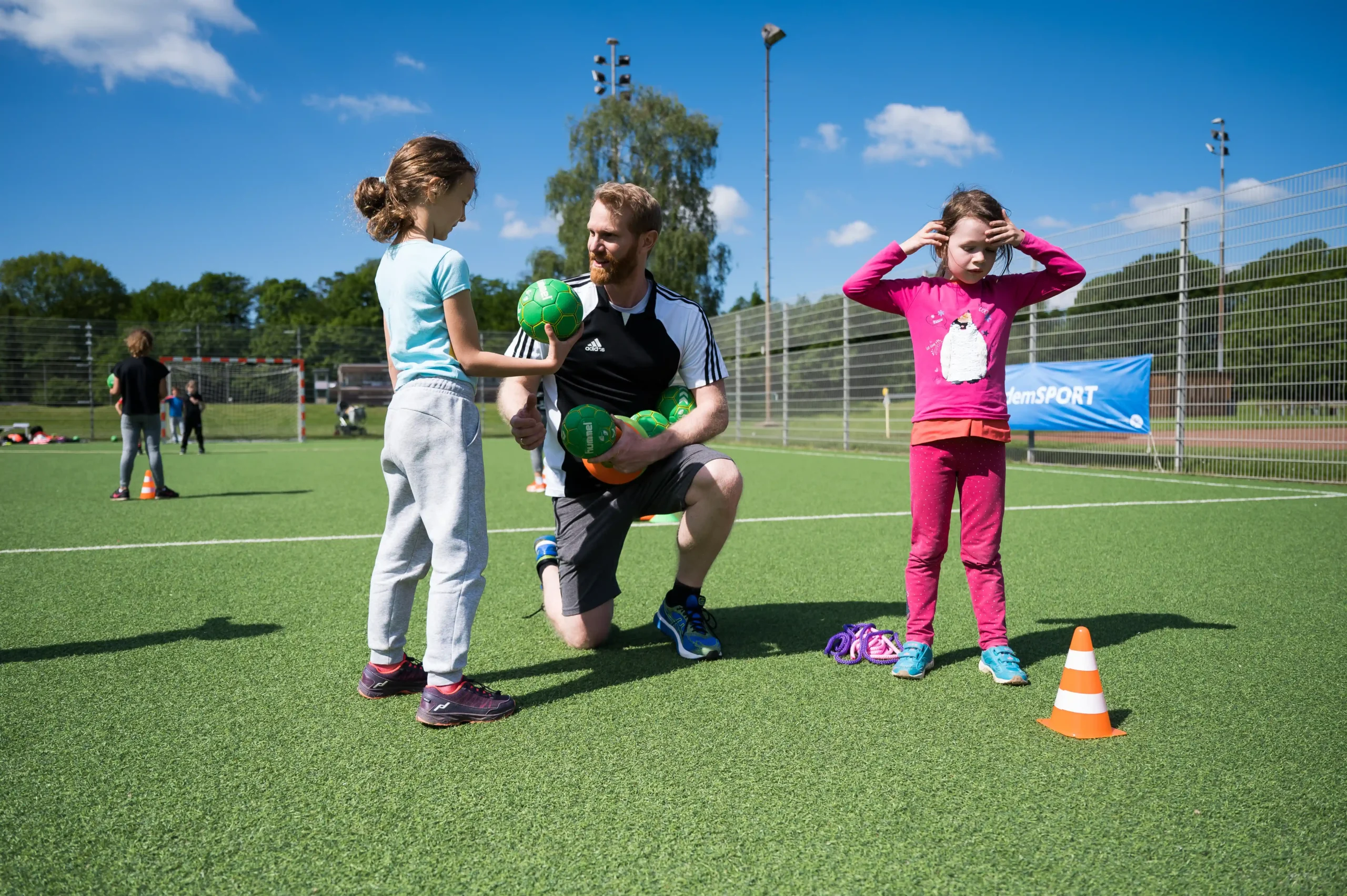 Ein Trainer kniet auf einem Sportplatz und reicht einem Mädchen einen grünen Ball, während ein anderes Mädchen daneben neben einem orangefarbenen Kegel steht.