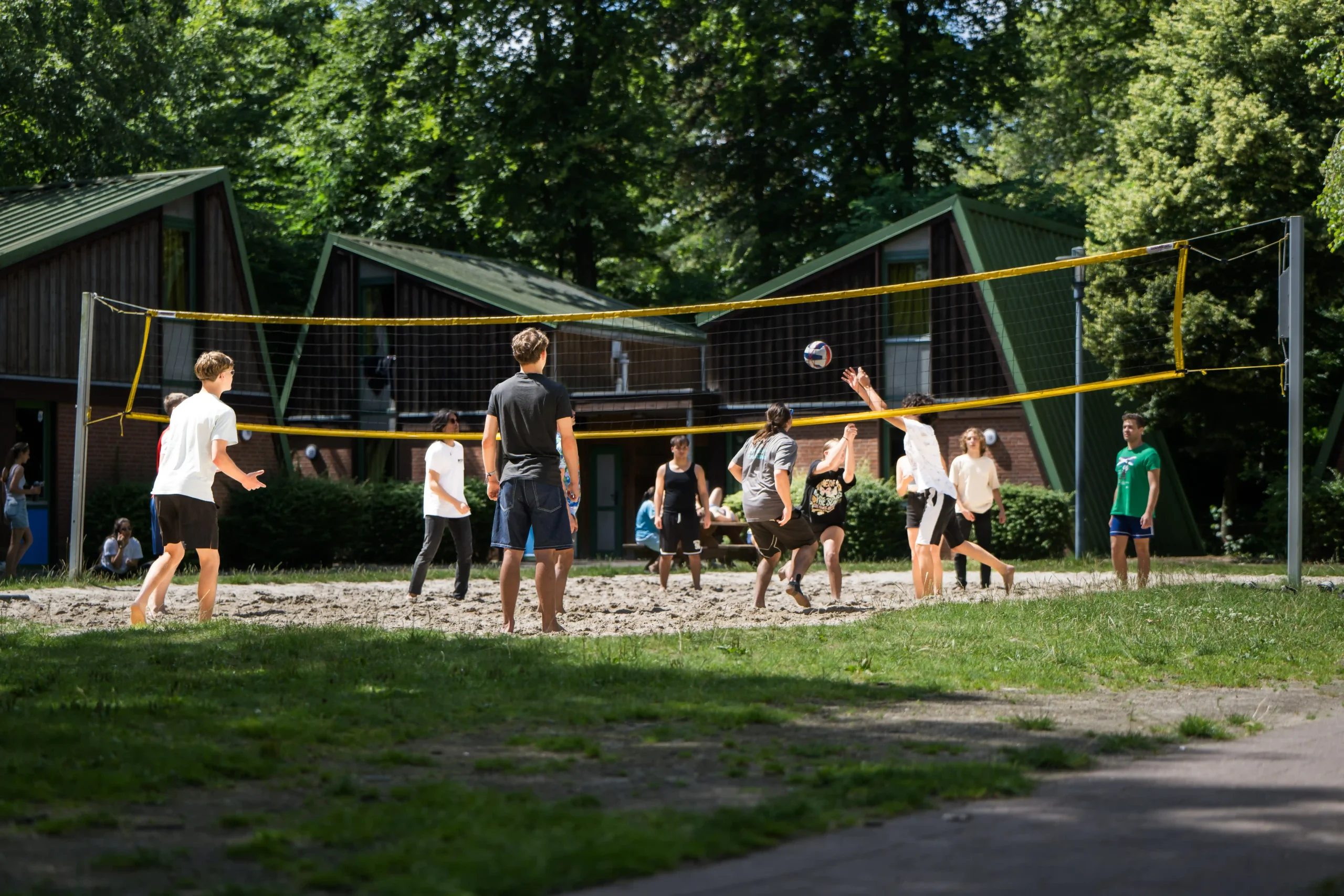 BSJ_Bremen_Volleyball_Jugendliche An einem sonnigen Tag spielt eine Gruppe von Menschen auf einem Sandplatz im Freien in der Nähe von Holzhütten, die von Bäumen umgeben sind, Volleyball.