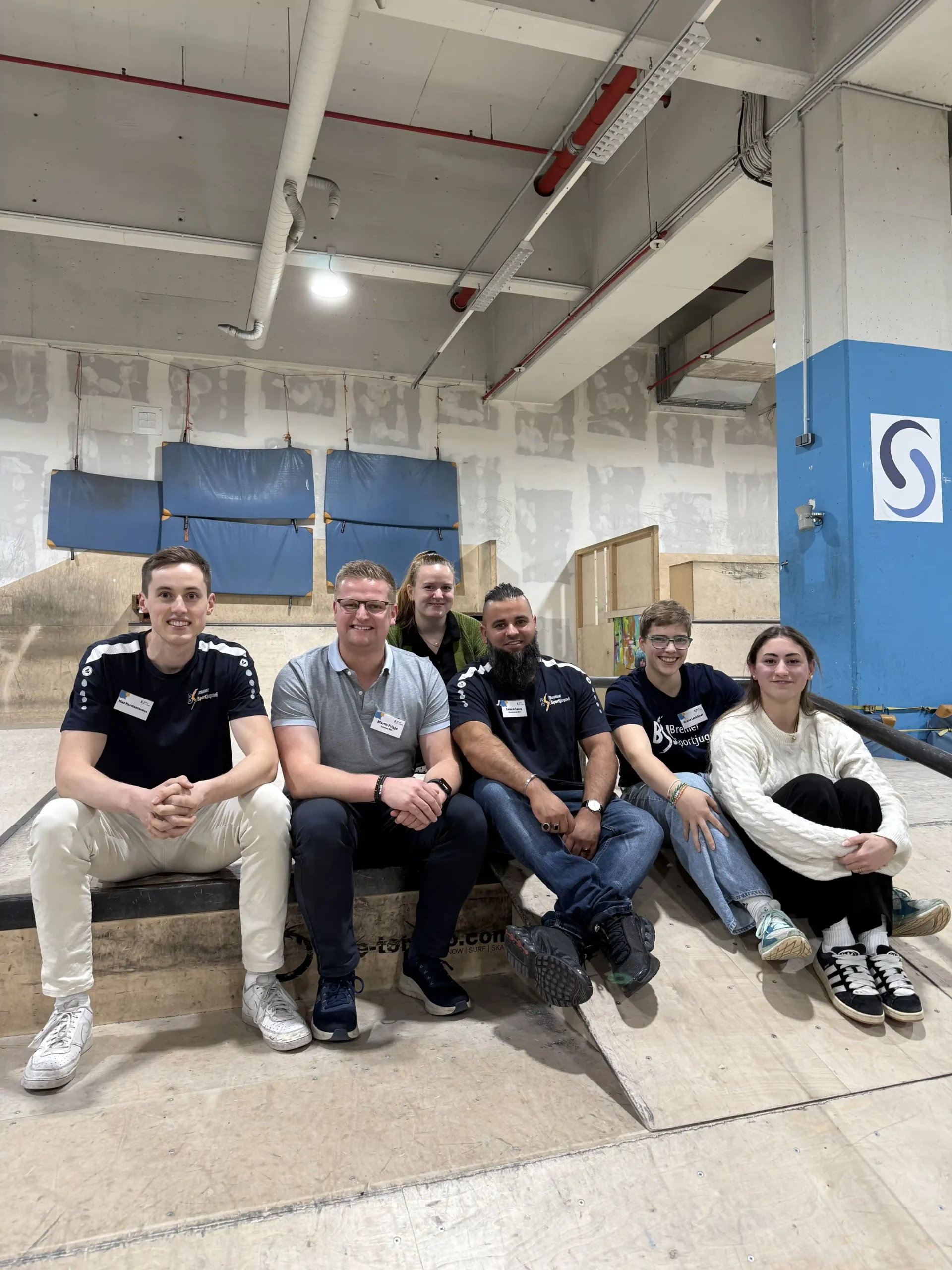 Sechs Personen sitzen nebeneinander auf einer Rampe in einem Indoor-Skatepark und lächeln in die Kamera. Im Hintergrund sind unfertige Wände und blaue Schutzpolster zu sehen.