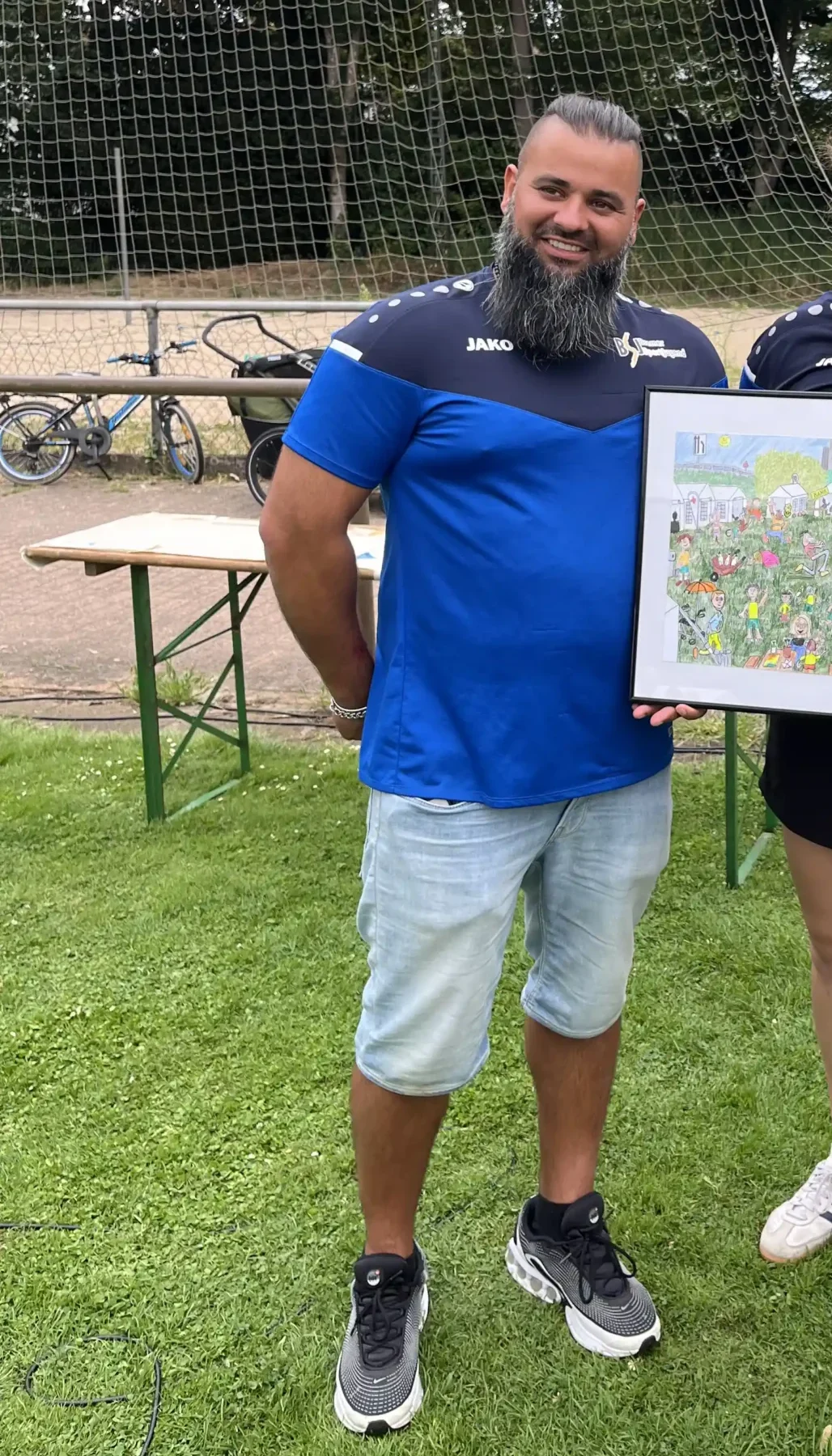 Eine Person in einem blauen Sportshirt und hellen Shorts steht auf dem Rasen neben einer anderen Person, die ein bunt gerahmtes Bild in der Hand hält. Im Hintergrund sind Fahrräder und ein Zaun zu sehen.