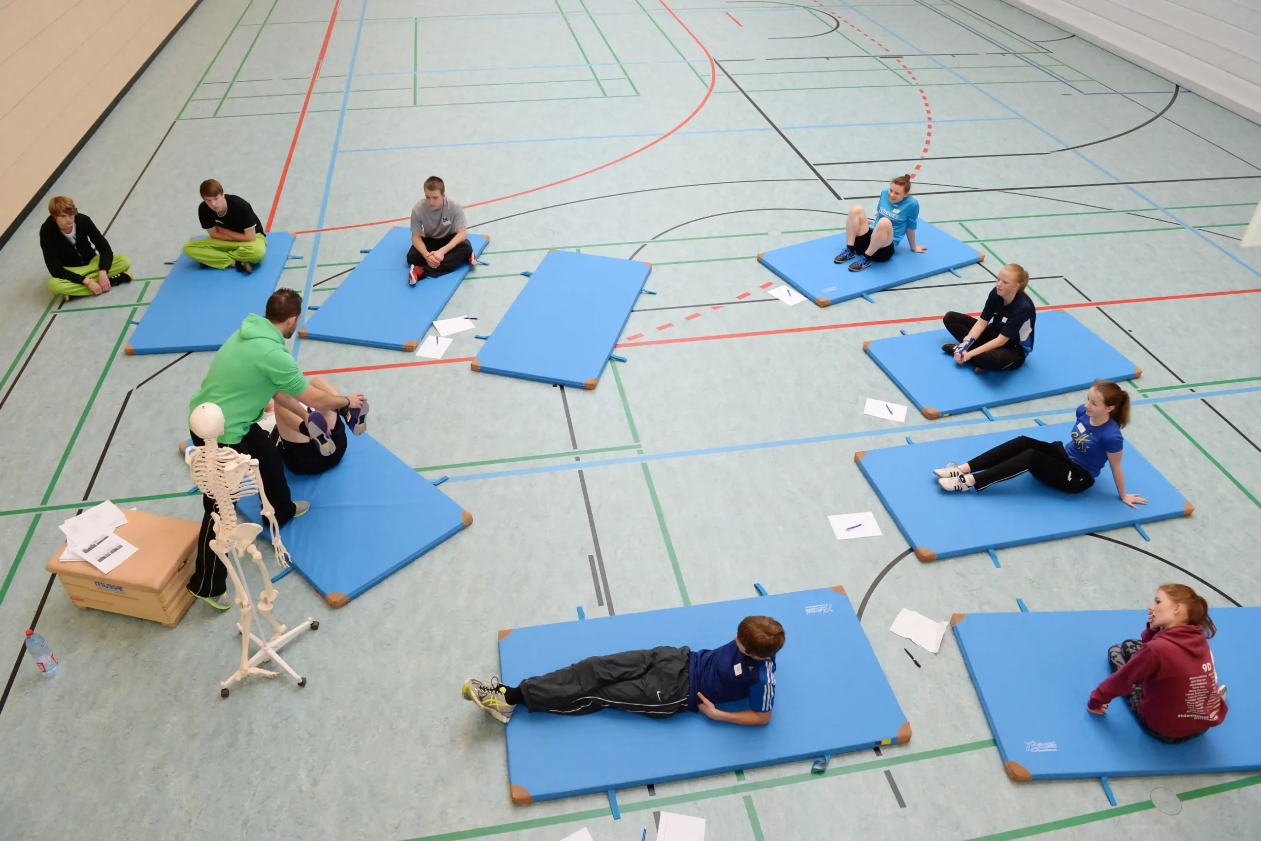 Acht Personen sitzen oder liegen auf blauen Gymnastikmatten, die in einer Turnhalle im Kreis ausgelegt sind; daneben liegen Papiere, und ein Skelettmodell ist zu sehen.
