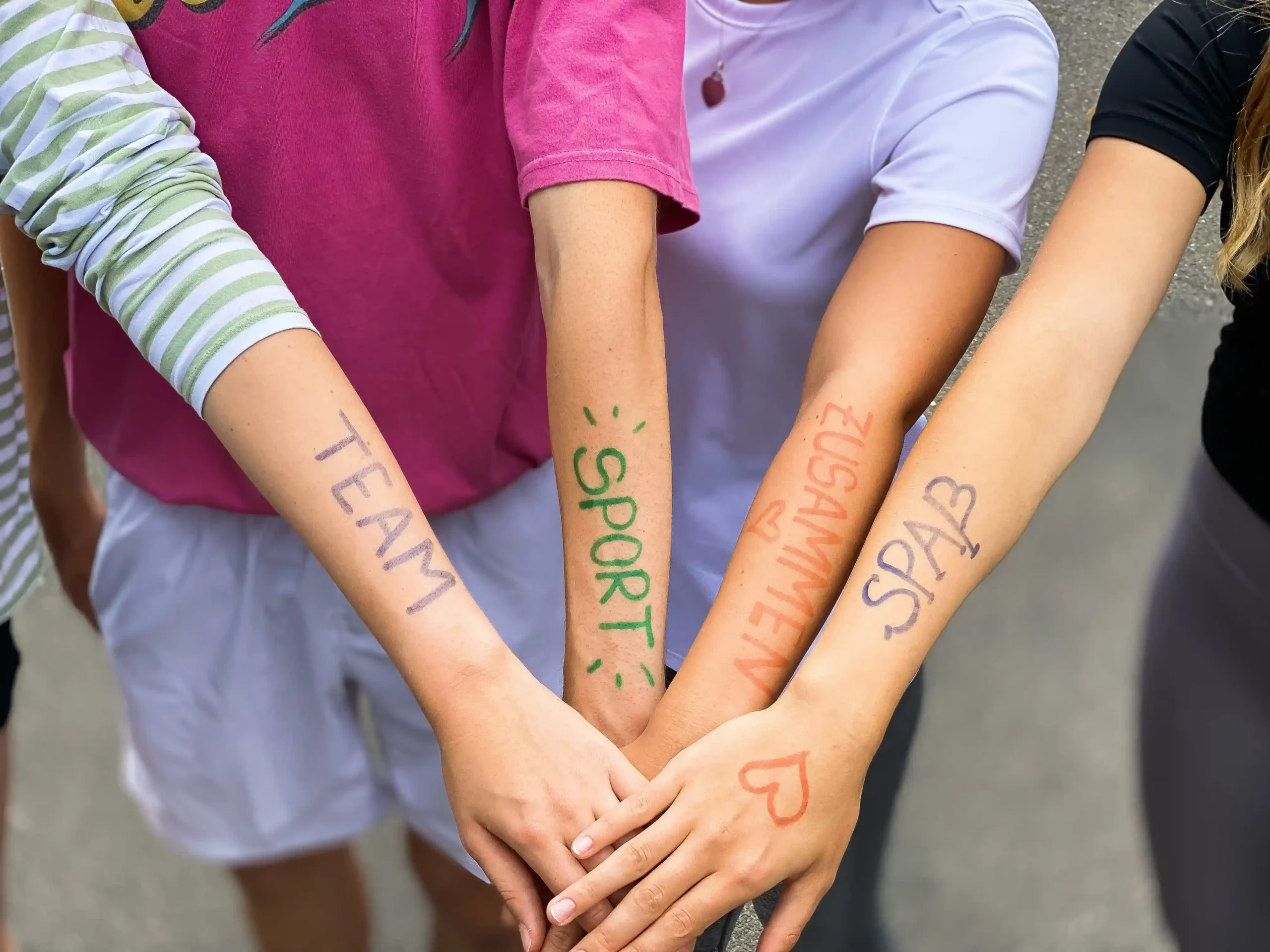 BSJ_Bremen_Zusammenhalt Vier Personen stehen in einer Reihe mit ausgestreckten Armen, auf jedem Unterarm sind mit bunten Filzstiften Wörter und Herzen aufgemalt: „TEAM“, „SPORT“, „ZUSAMMEN“ und „SPASS“.