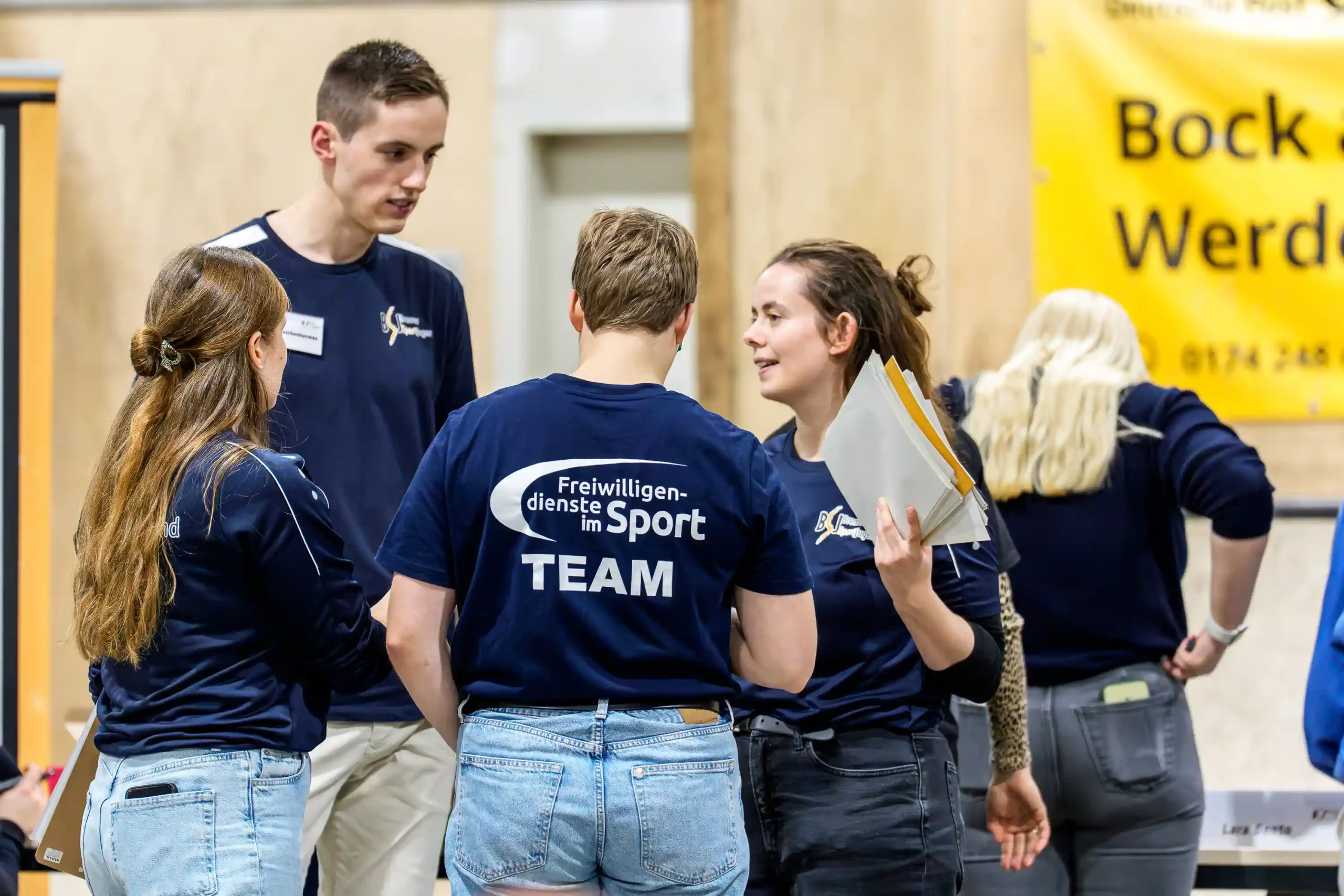 Vier Personen in einheitlichen marineblauen Shirts mit der Aufschrift „TEAM“ unterhalten sich in einem Raum, wobei zwei von ihnen Zettel in der Hand halten. Im Hintergrund hängt ein gelbes Transparent.