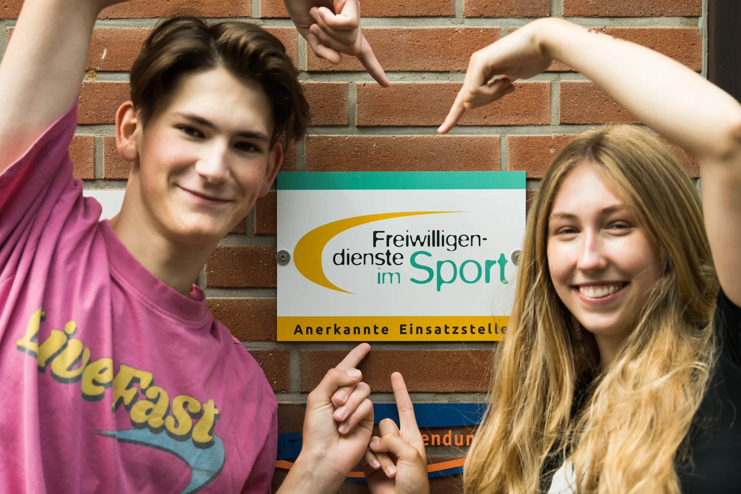 BSJ_FWD_im_Sport Zwei lächelnde junge Leute zeigen auf ein Schild an einer Backsteinmauer, auf dem „Freiwilligendienste im Sport – Anerkannte Einsatzstelle“ steht.
