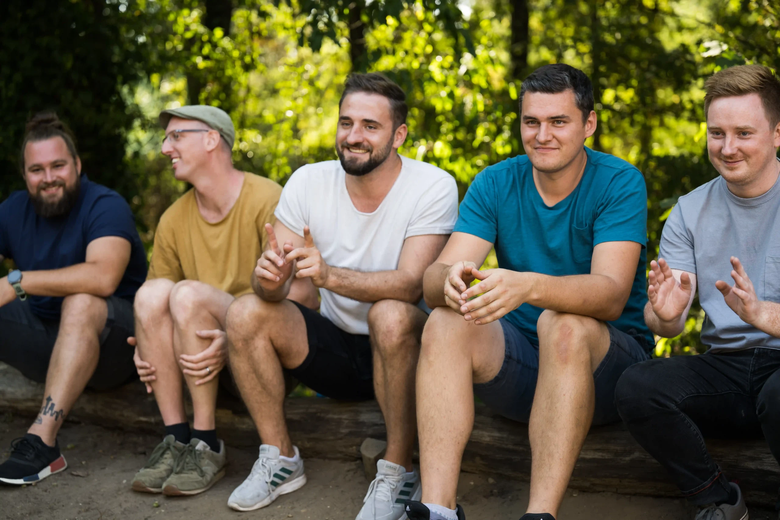BSJ_FWD_im_Sport_Bremen Fünf Männer sitzen draußen auf einem Baumstamm, leger gekleidet, lächelnd und klatschend, vor dem Hintergrund von grünem Laub.