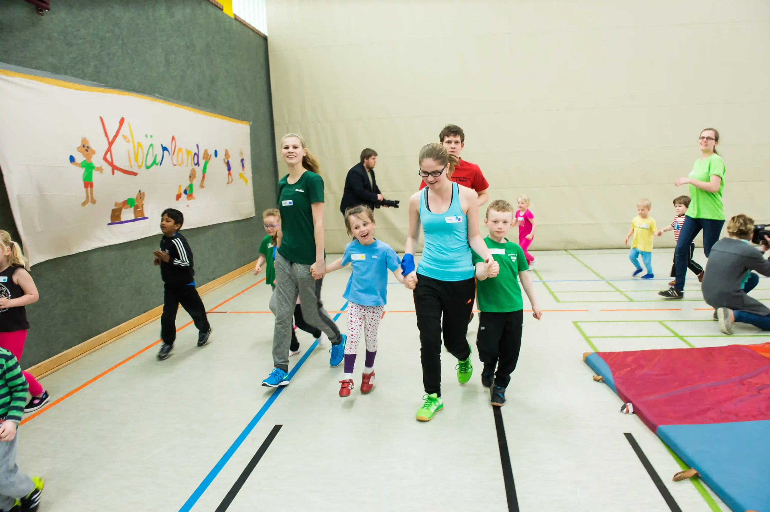 Erwachsene und Kinder nehmen an einer Gruppenaktivität in einer Turnhalle teil, gehen spazieren und spielen gemeinsam. Im Hintergrund hängt ein buntes Transparent an der Wand.