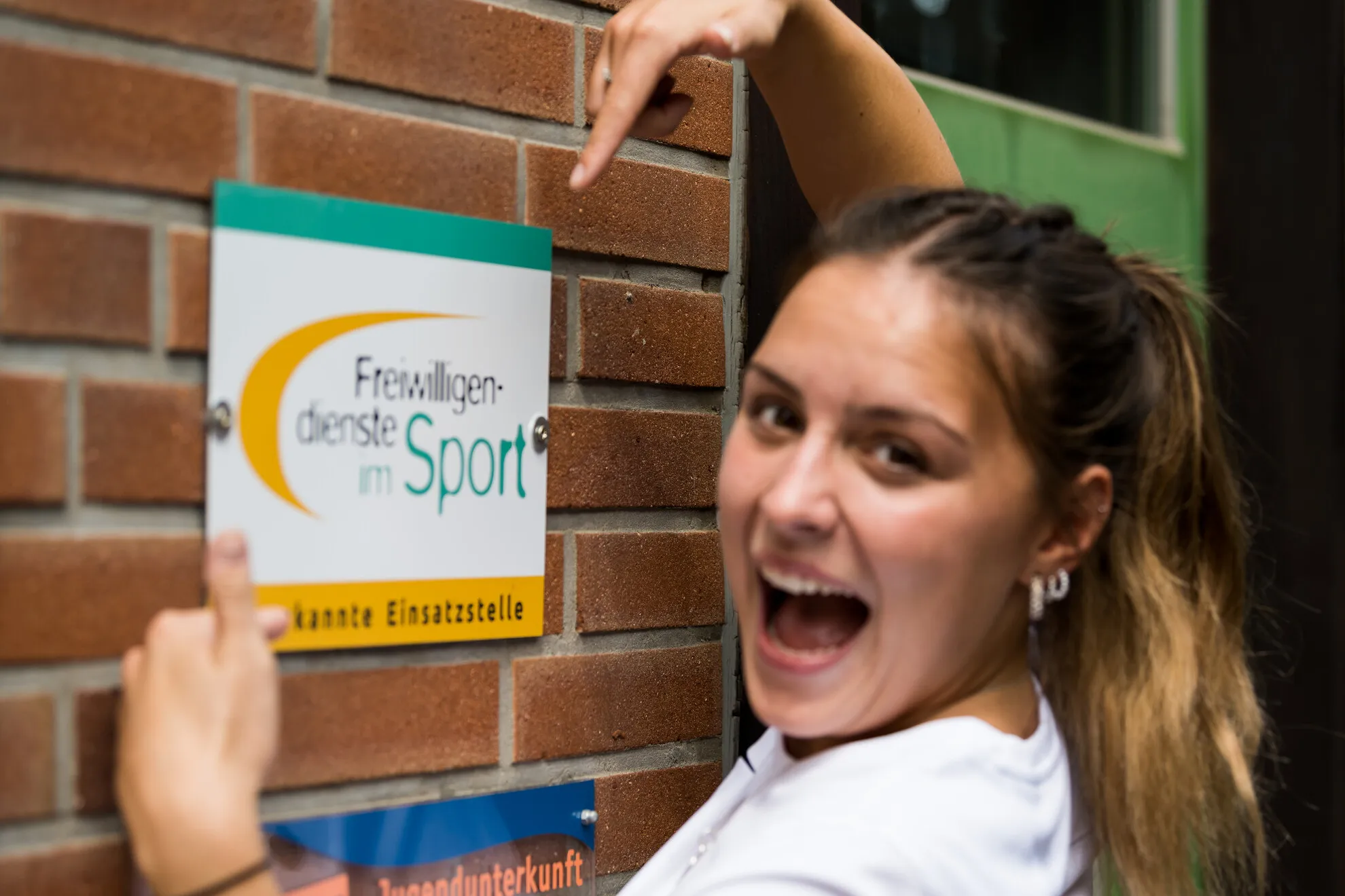 BSJ_Freiwilligendienste_im-Sport Eine lächelnde Person zeigt aufgeregt auf ein Schild an einer Backsteinmauer, auf dem „Freiwilligendienste im Sport – anerkannte Einsatzstelle“ steht.