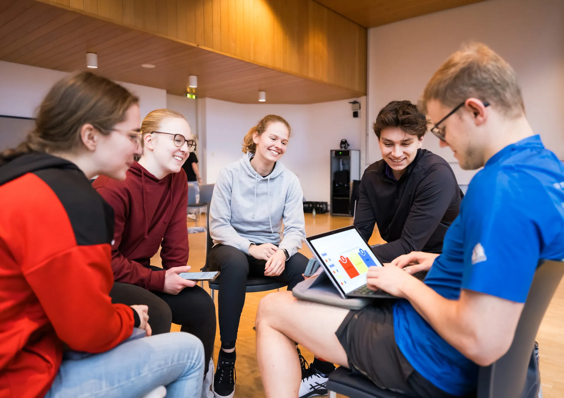 Fünf junge Erwachsene sitzen in einem Raum im Kreis, wobei eine Person der Gruppe auf einem Laptop ein Diagramm zeigt. Sie scheinen sich in einer Diskussion zu befinden.