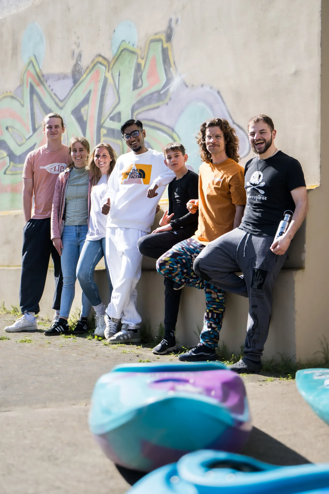 Sieben Menschen stehen im Freien Seite an Seite vor einer Graffiti-Wand, posieren und lächeln, während im Vordergrund bunte Kajaks auf dem Boden liegen.