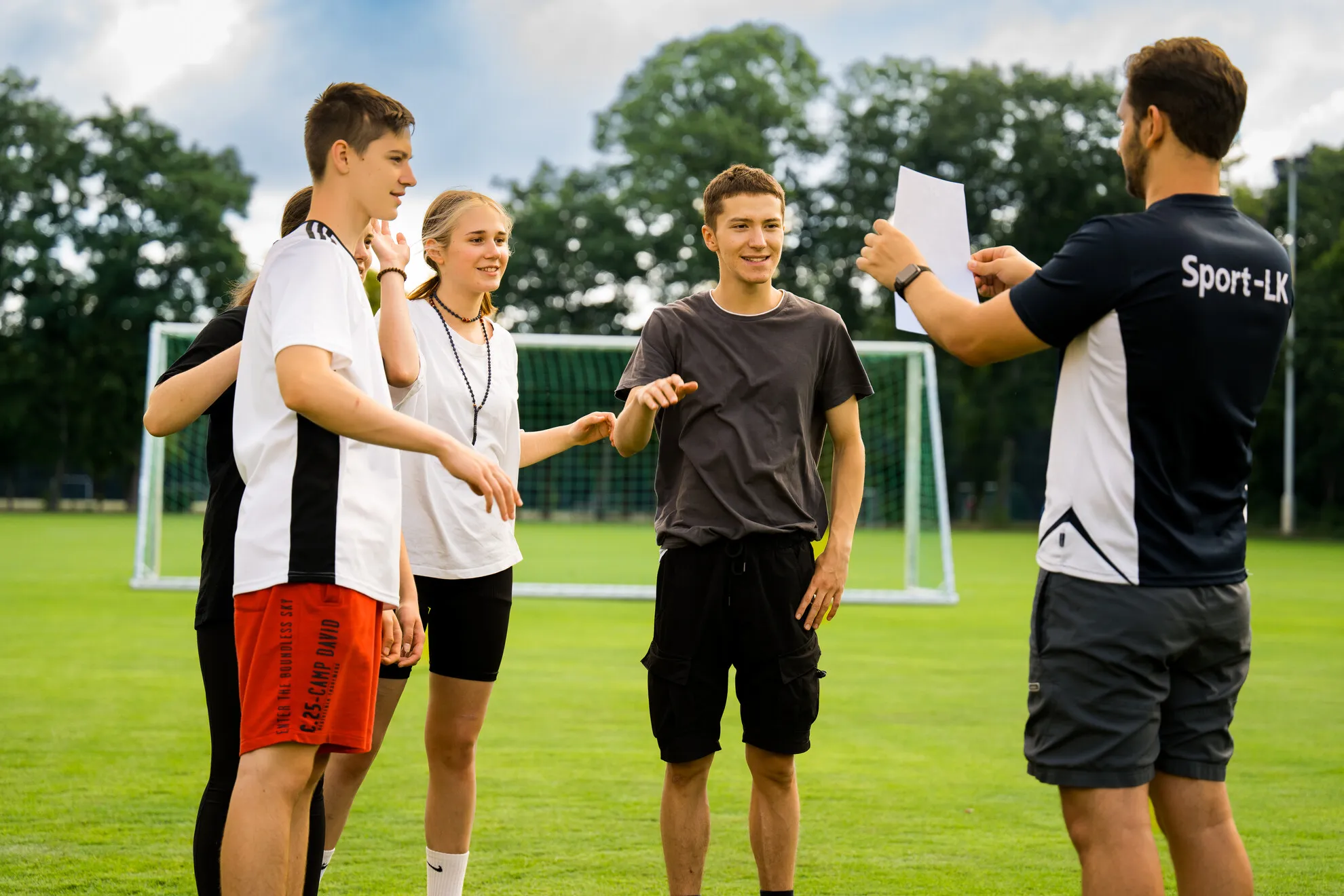 BSJ_Jugendbildung_Team_junge-Menschen Ein Trainer zeigt einen Zettel an vier Jugendliche, die auf einem Fußballplatz stehen, im Hintergrund sind Torpfosten und Bäume zu sehen.