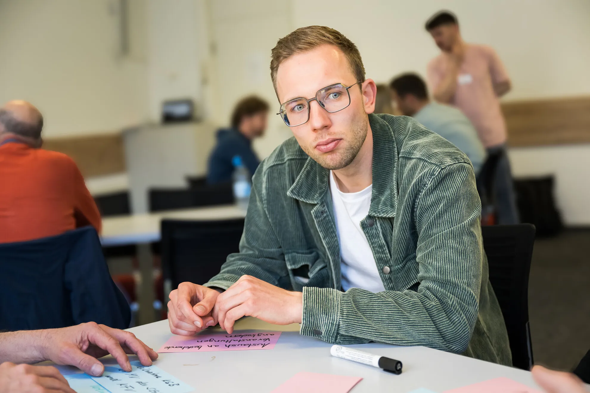 Ein Mann mit Brille und grüner Jacke sitzt mit anderen an einem Tisch und hält während einer Gruppenaktivität in einem Klassenzimmer Stift und Papier in der Hand.