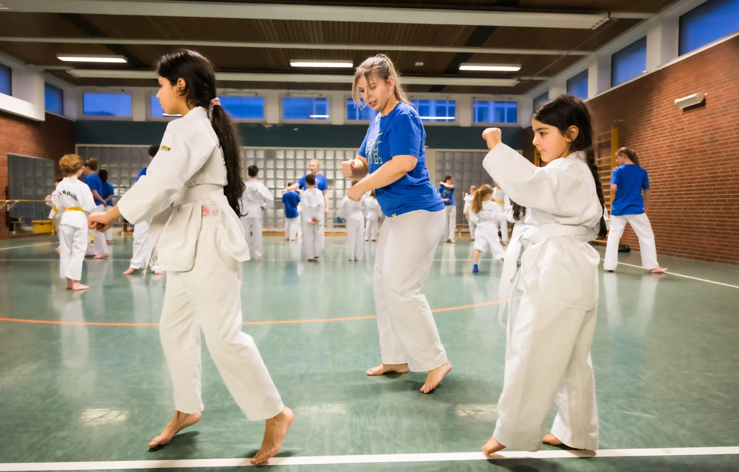 Eine Gruppe von Kindern und ein Erwachsener üben in einer Turnhalle Kampfsportübungen; alle tragen weiße Kampfsportanzüge und stehen barfuß auf einem grünen Boden.