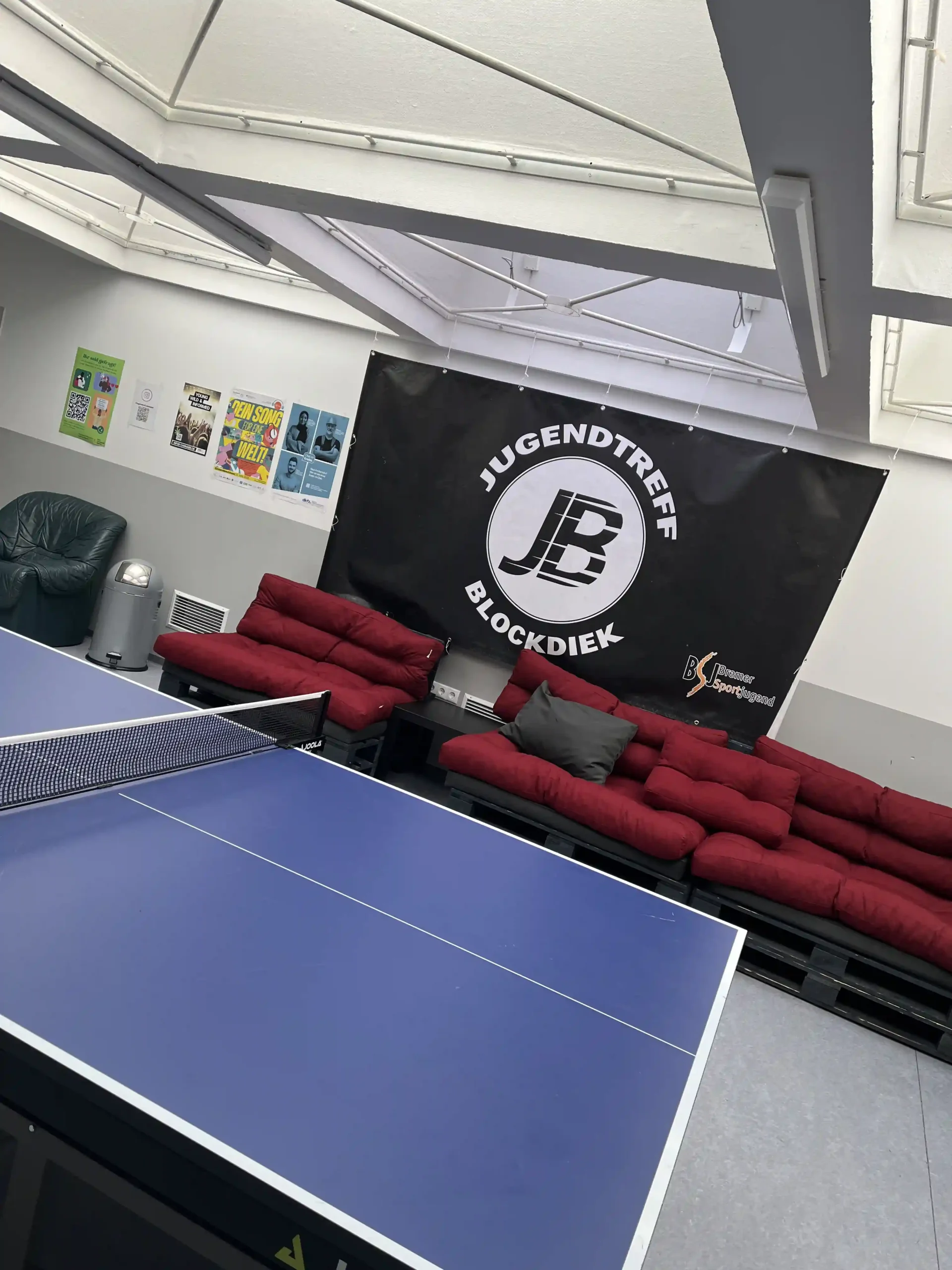 Ein Tischtennistisch vor roten Sofas und ein großes schwarzes Banner mit der Aufschrift „Jugendtreff Blockdiek“ in einem Raum, dessen Wände mit Plakaten geschmückt sind.